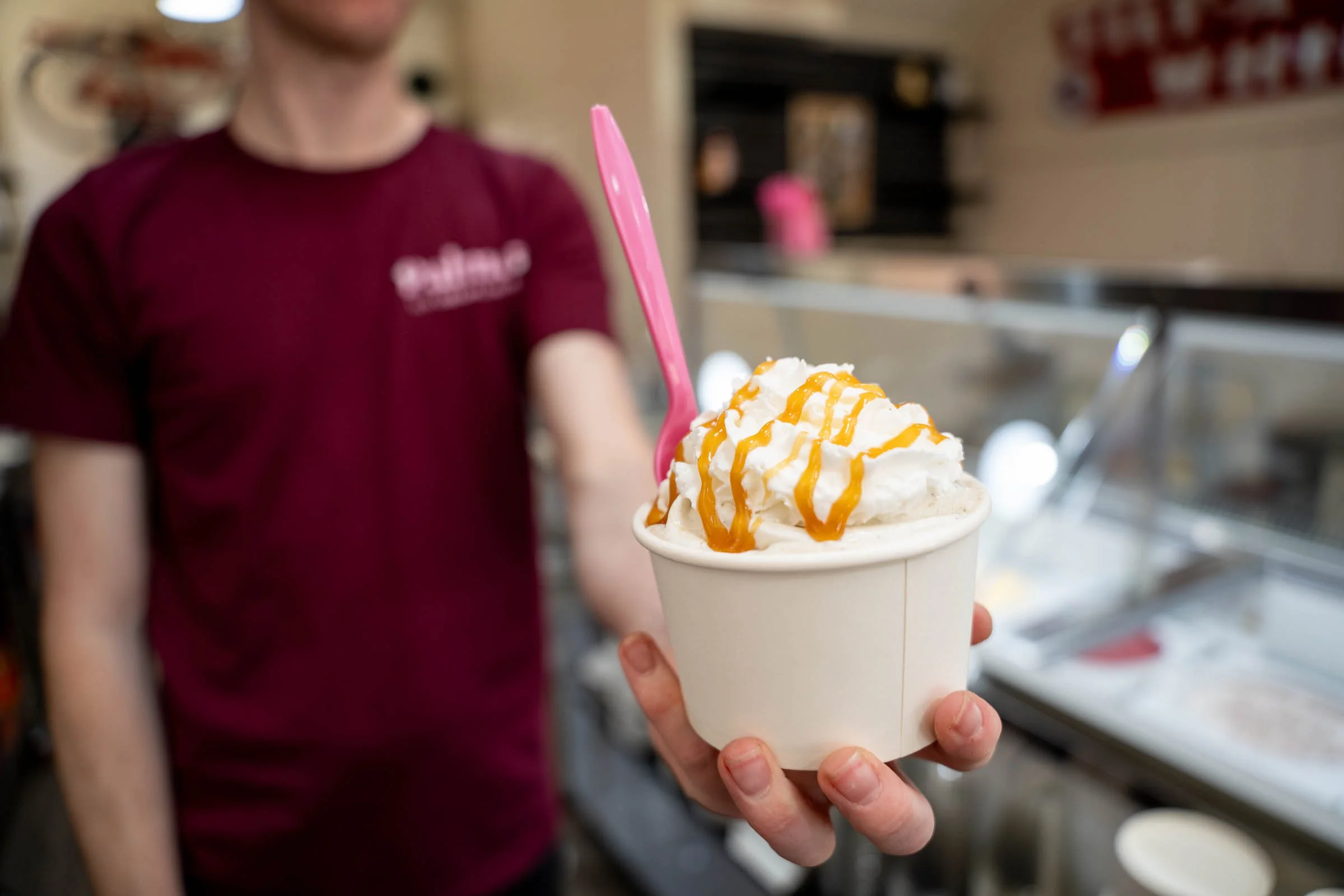 A person holding a cup of vanilla soft serve ice cream topped with caramel drizzle and whipped cream, with a pink spoon inserted, in a casual setting.