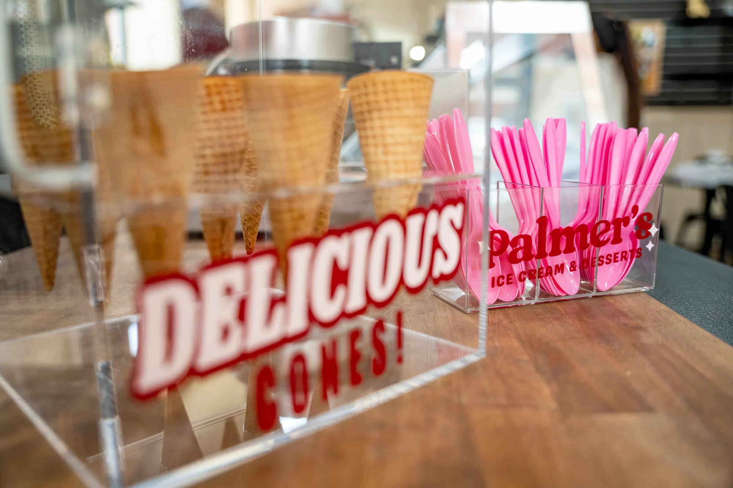 Pink plastic spoons in a clear container labeled "Palmers Ice Cream & Desserts" on a wooden counter, with waffle cones and a coffee machine in the background.