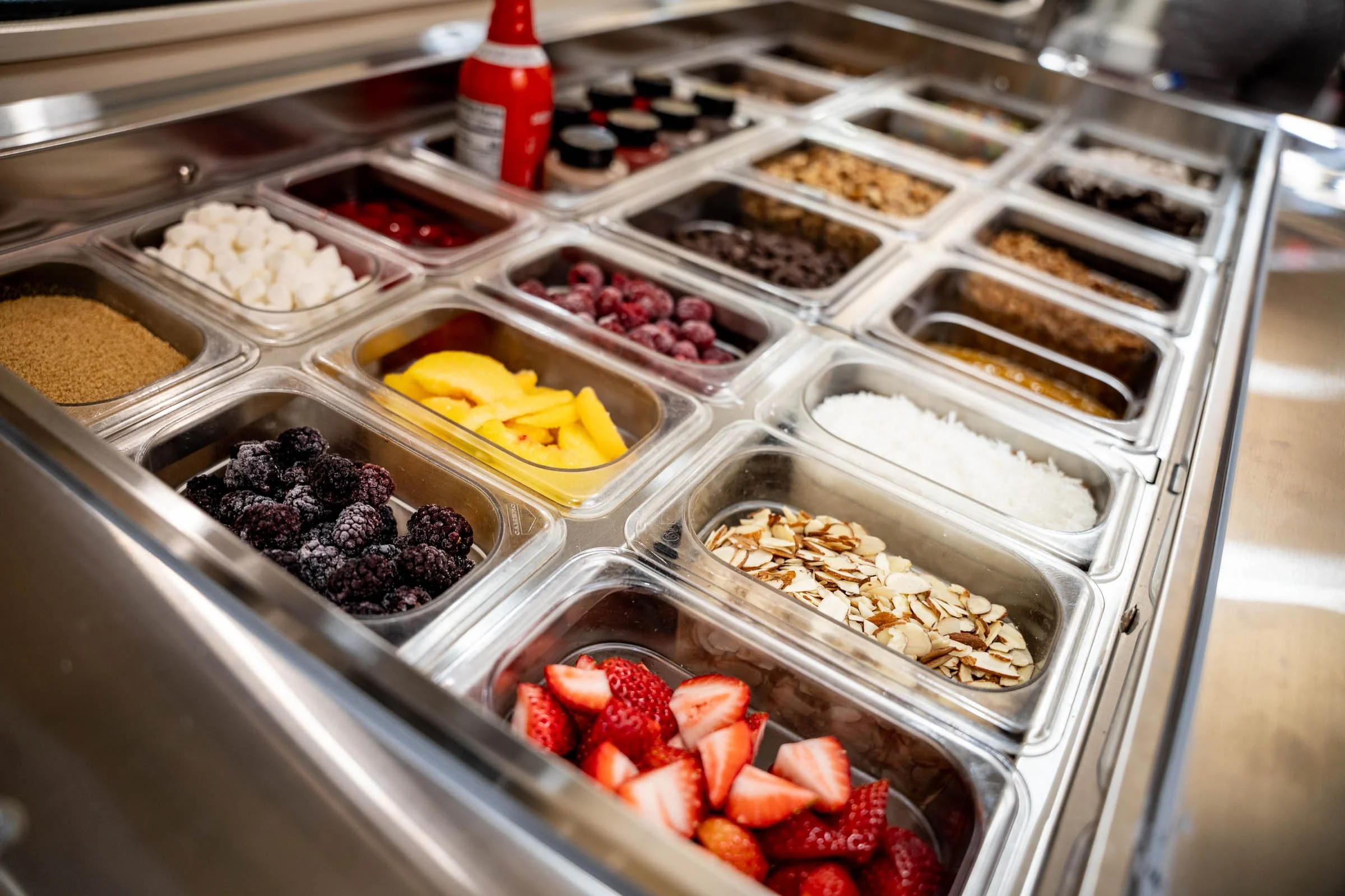A display case of various toppings for ice cream, including fresh strawberries, blackberries, sliced almonds, shredded coconut, fruit syrups, candies, and pieces of pineapple and other fruits.