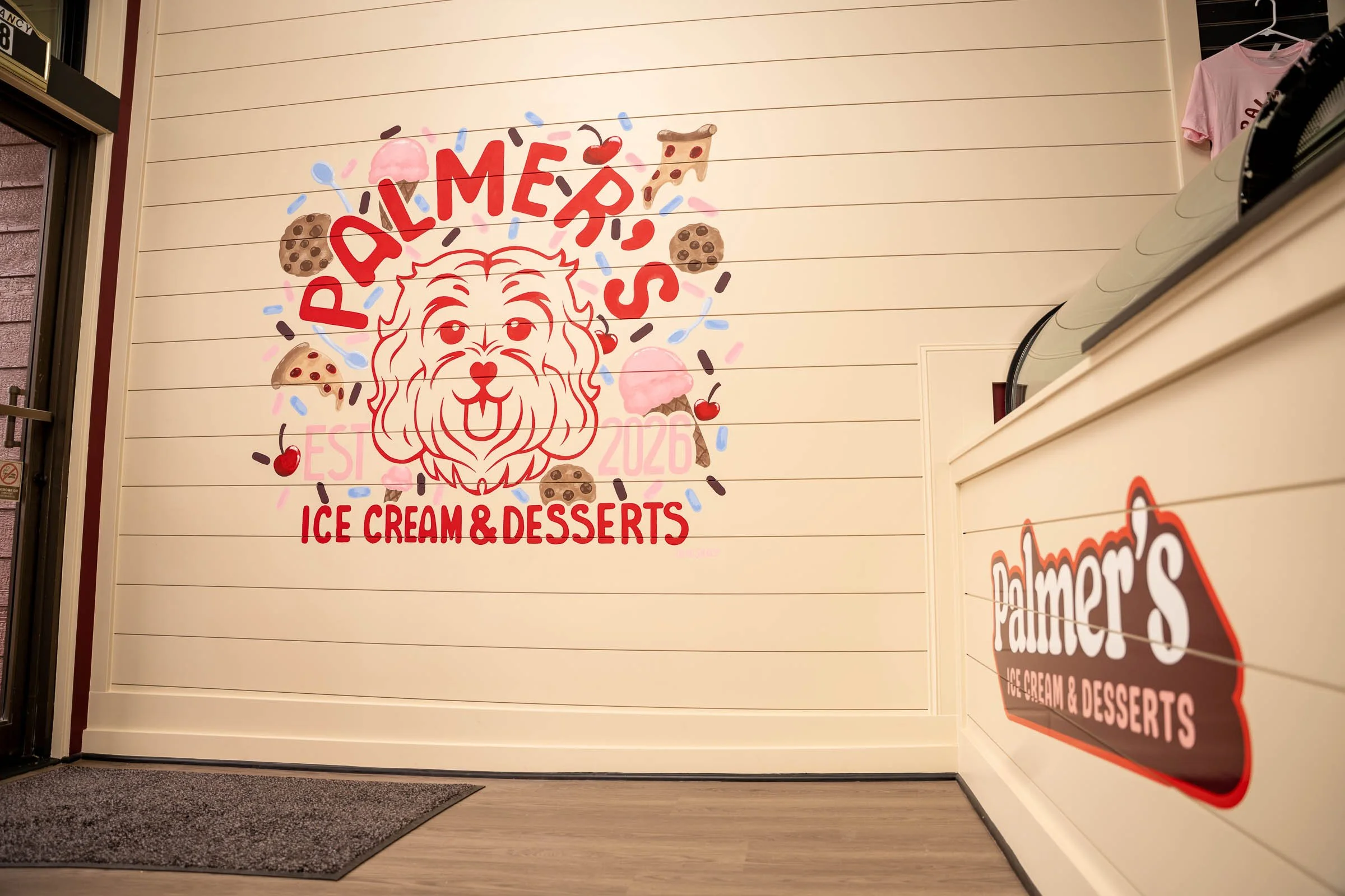 Sign that reads 'Palmer's Ice Cream & Desserts' with a dog face illustration, ice cream cones, slices of pizza, and cherries around it, on a cream-colored wall in a store.