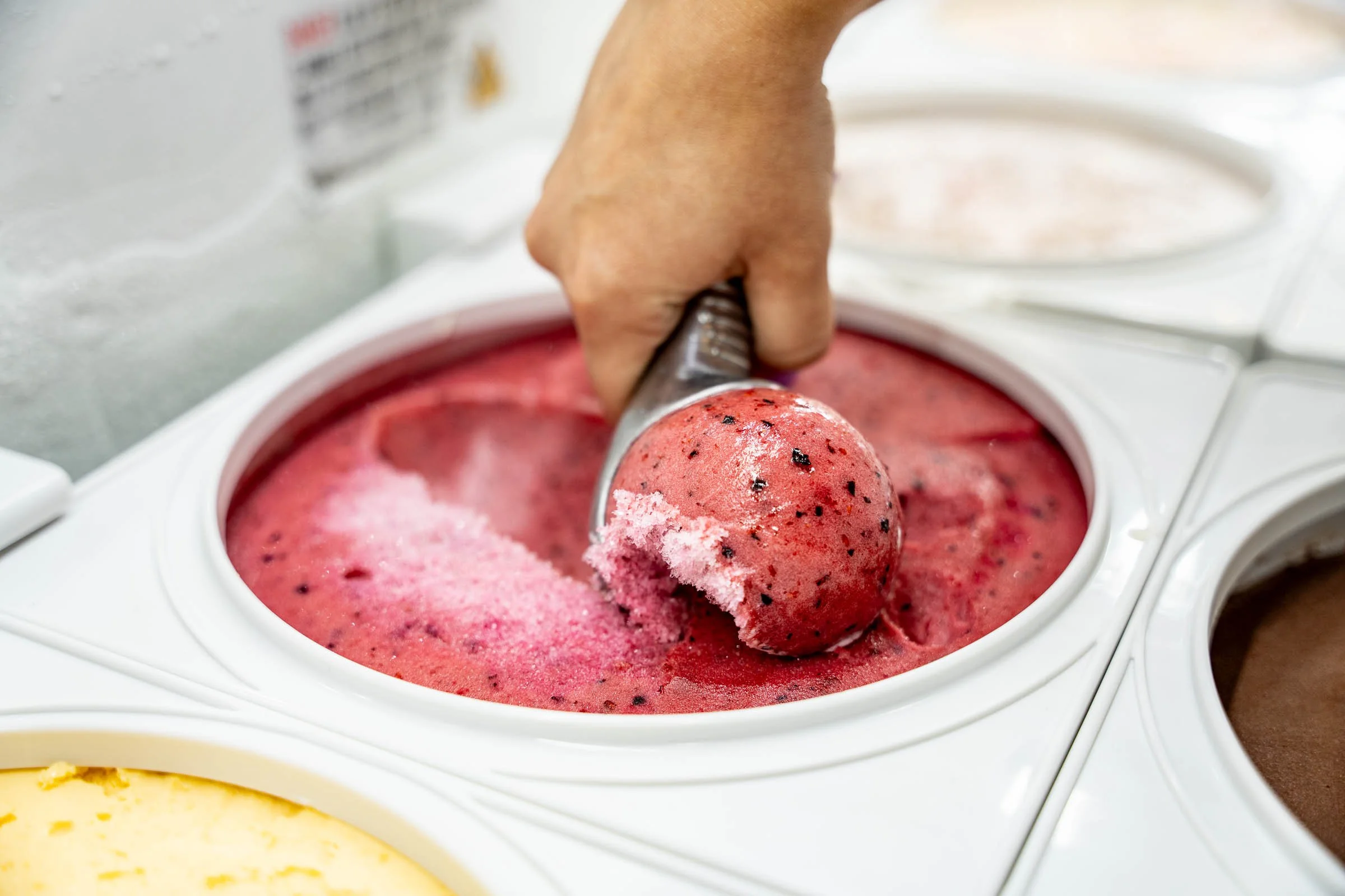 Close-up of a hand scooping pink berry ice cream from an ice cream maker with multiple flavors visible.