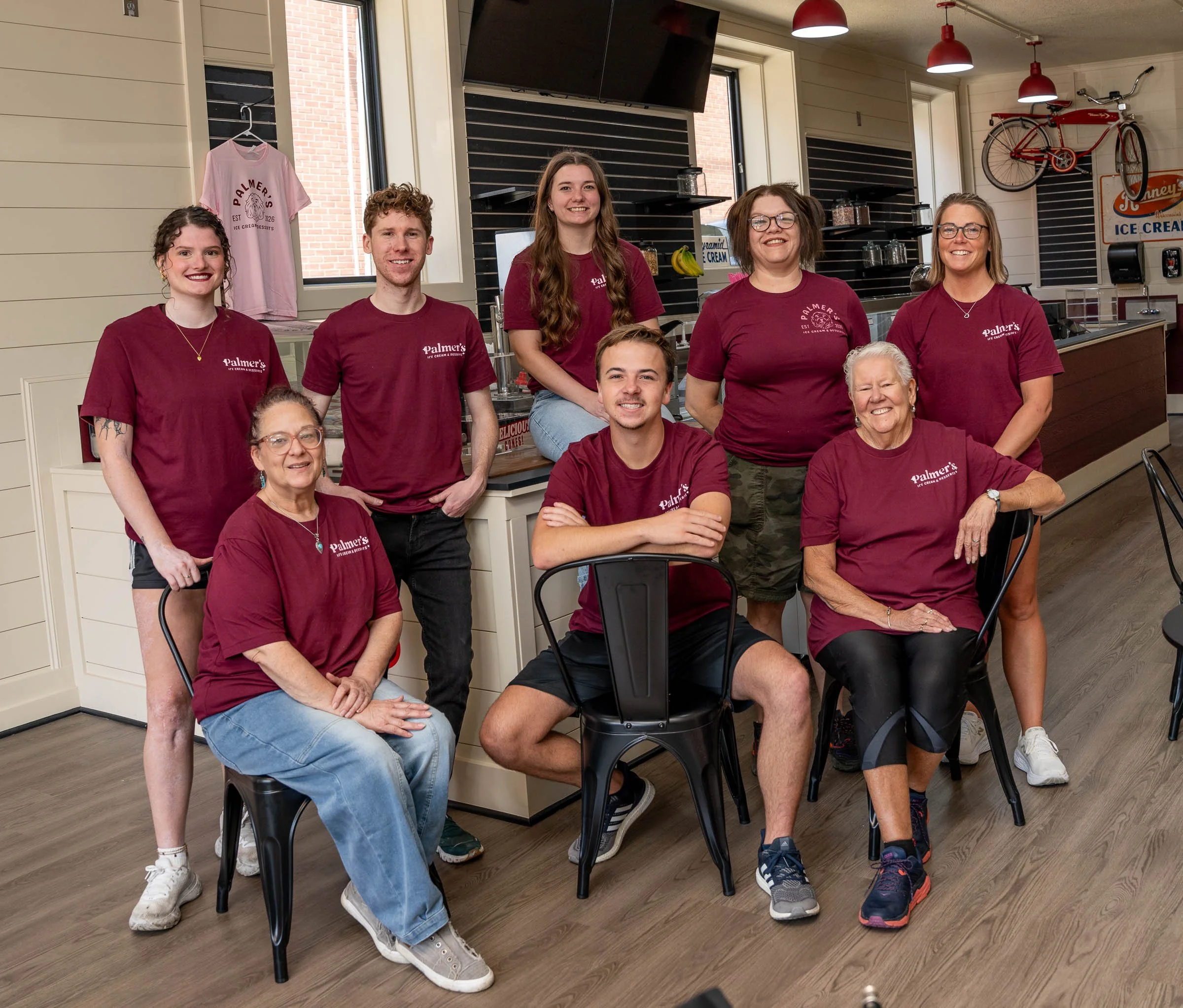 A group of nine people, six women and three men, wearing maroon t-shirts with 'Palmer's Ice Cream' logo, posing in a casual indoor setting at an ice cream shop or cafe, smiling at the camera.