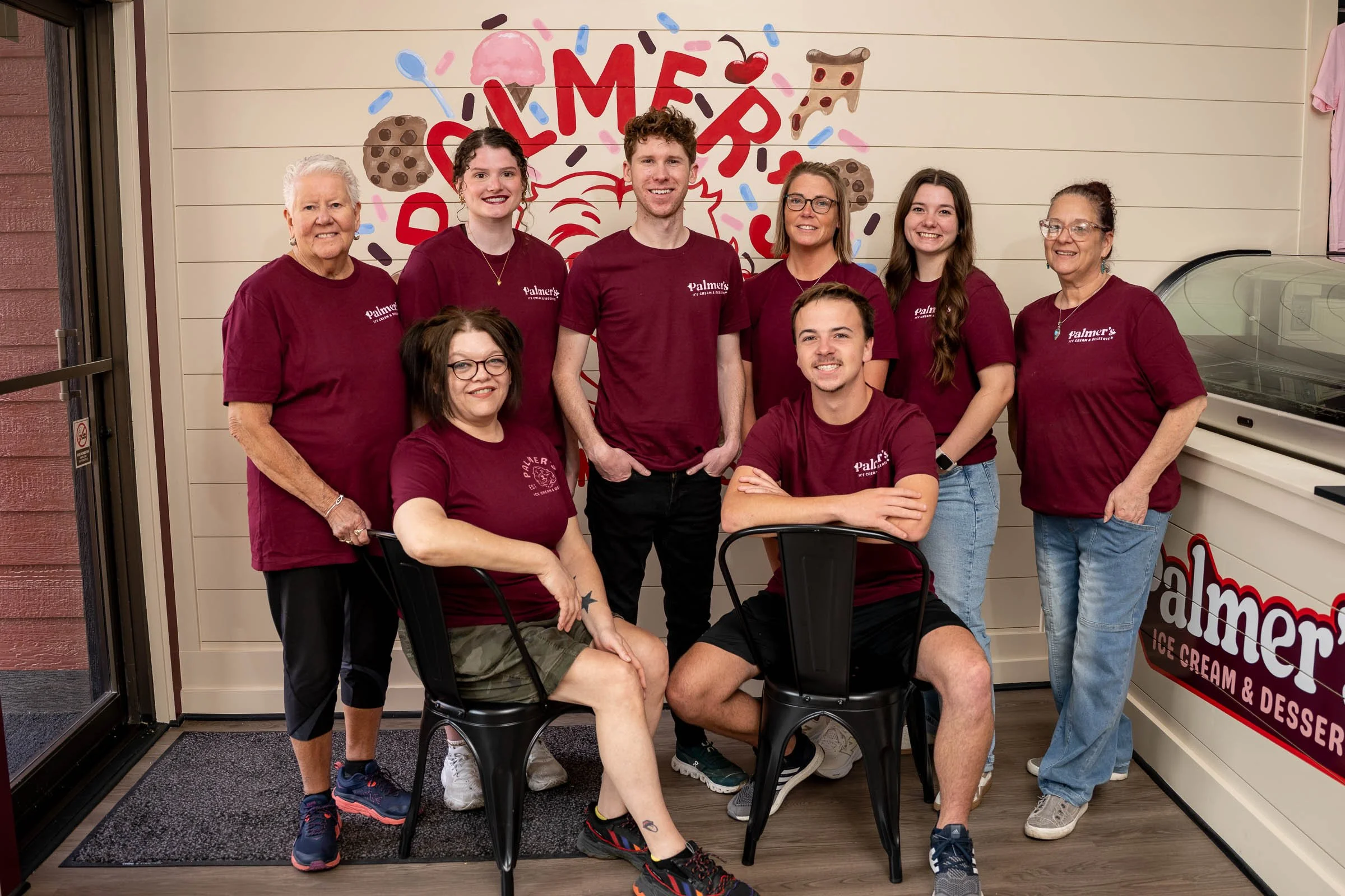 Group of eight people posing inside an ice cream shop, standing in front of a colorful mural with ice cream and cookie images. They are all wearing maroon shirts with the shop's logo. Two women are seated on black chairs, while the others stand behind them.