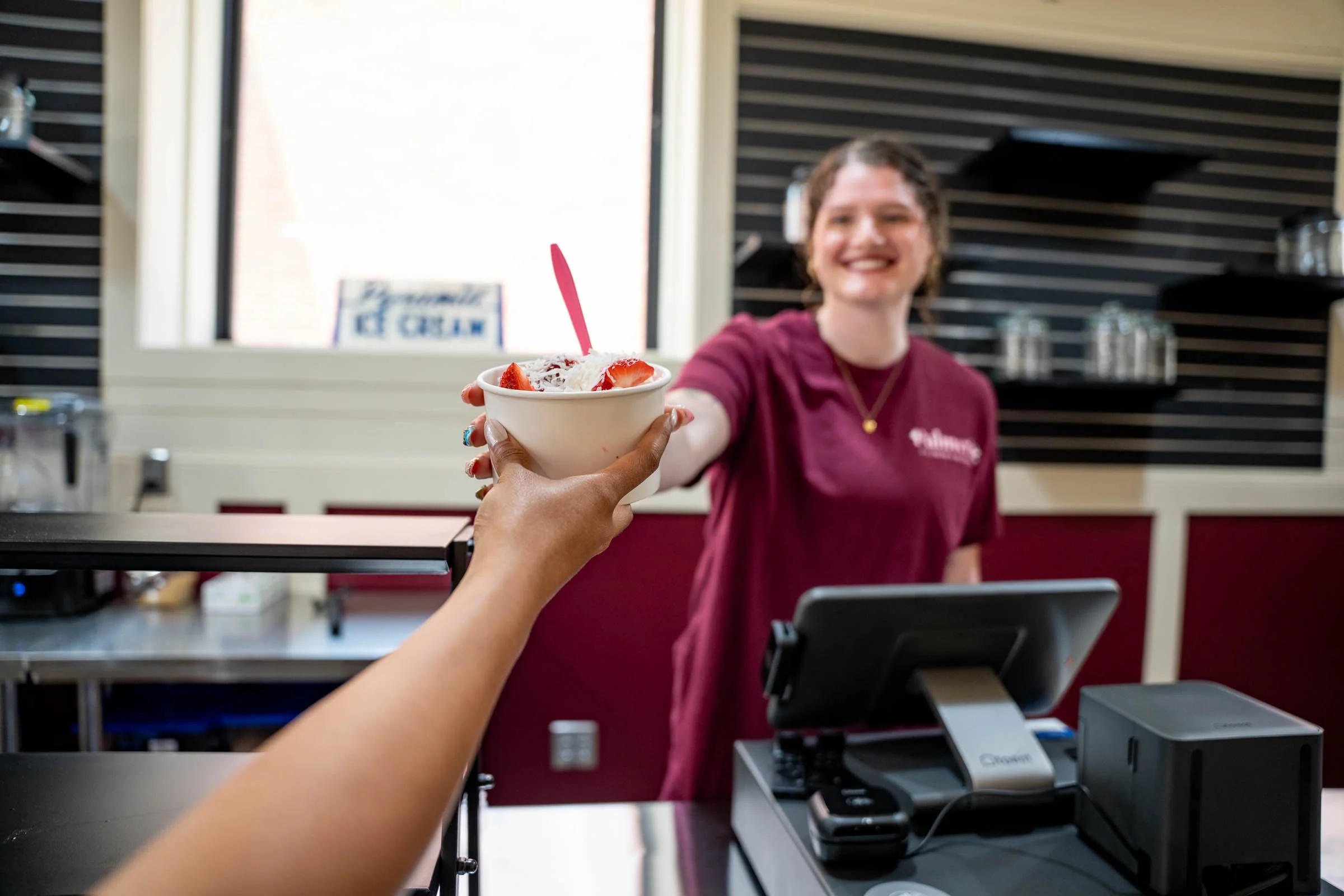 A person is holding a bowl of ice cream with strawberries, whipped cream, and a pink spoon towards a smiling woman behind a counter in an ice cream shop.