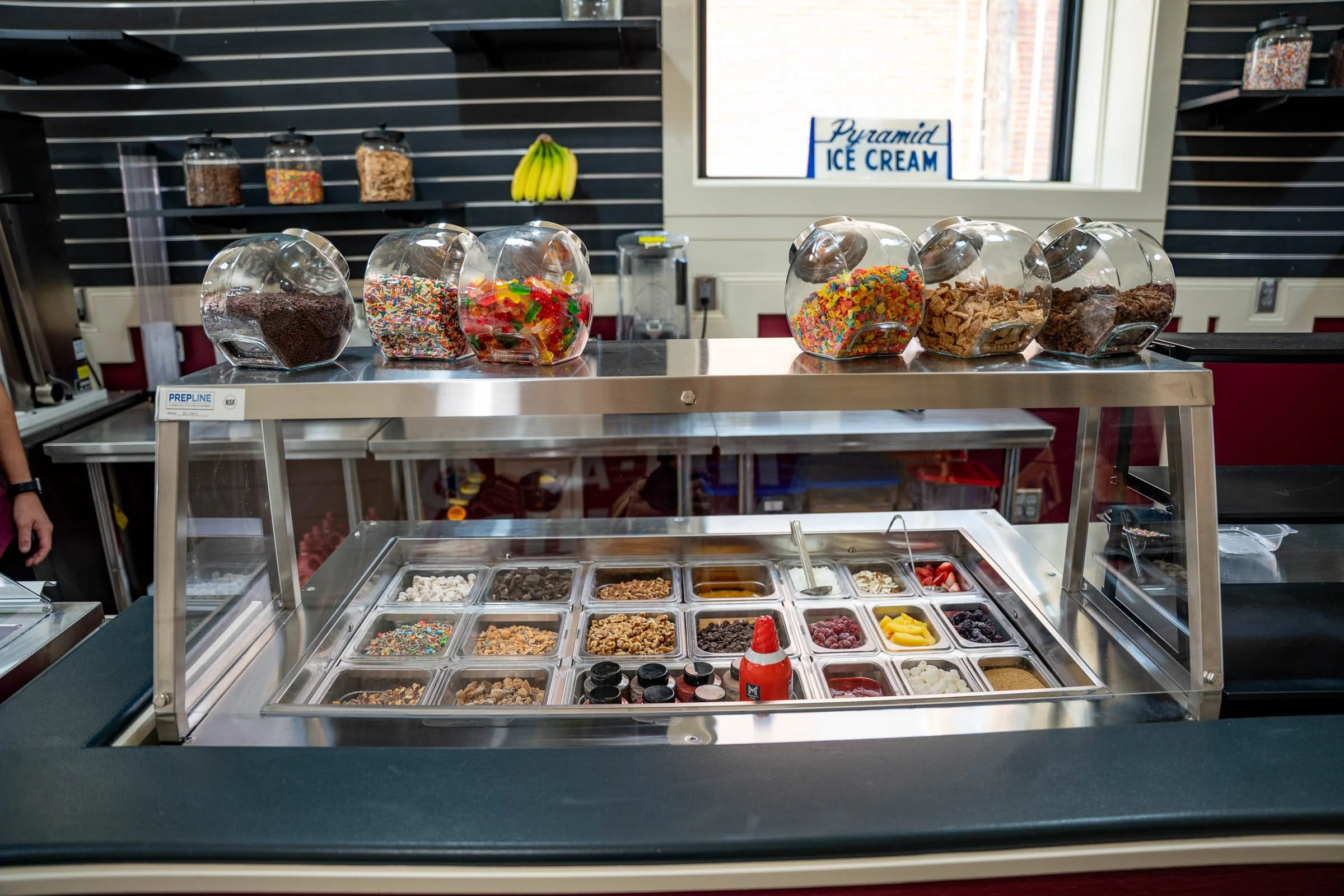 Ice cream shop with various toppings and mix-ins in glass containers, and a sign reading 'Pyramid Ice Cream' in the background.