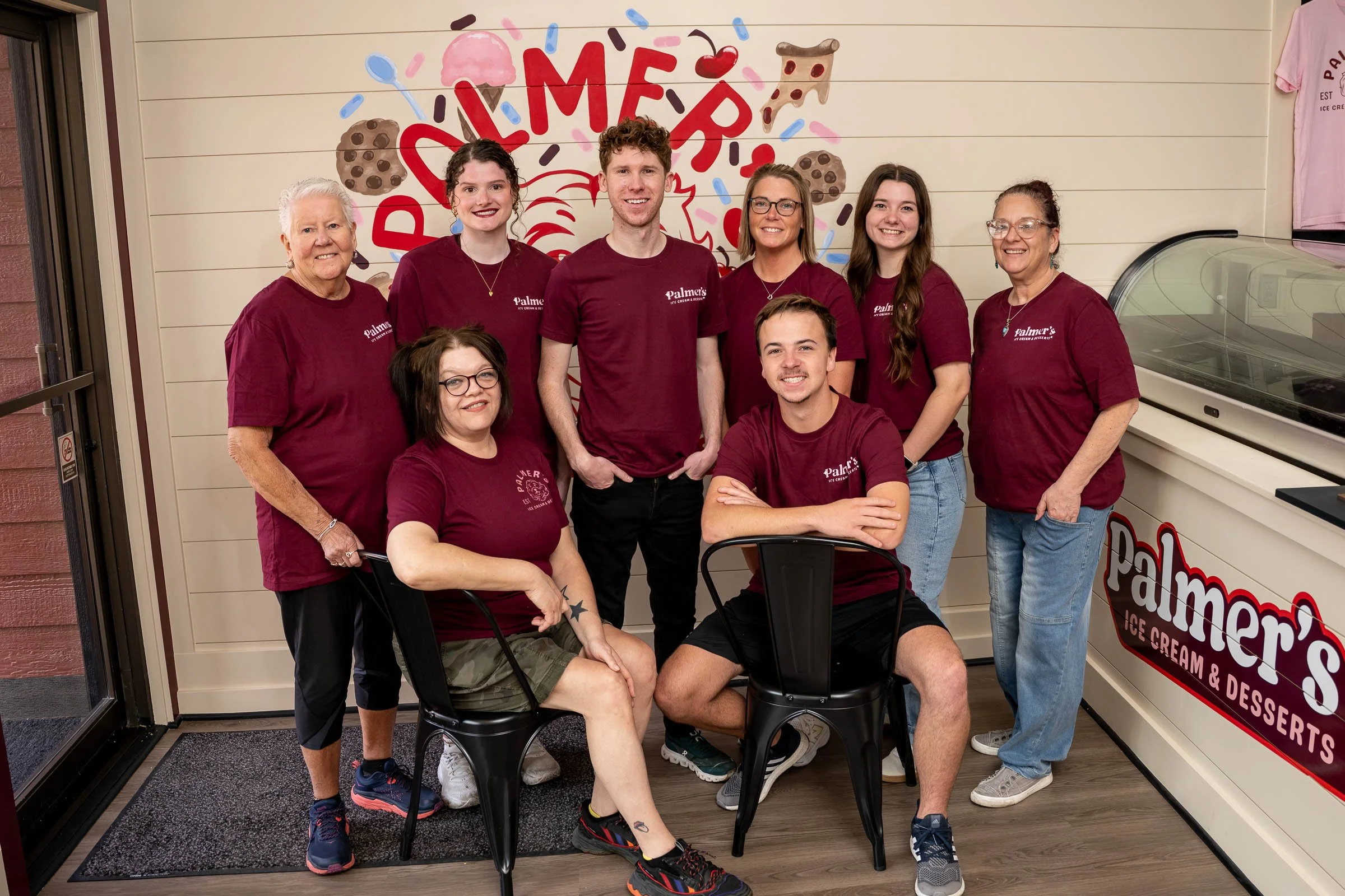 Group of seven people standing and sitting in front of an ice cream shop, smiling and wearing matching maroon shirts with the shop's logo, with colorful ice cream and dessert illustrations on the wall behind them.