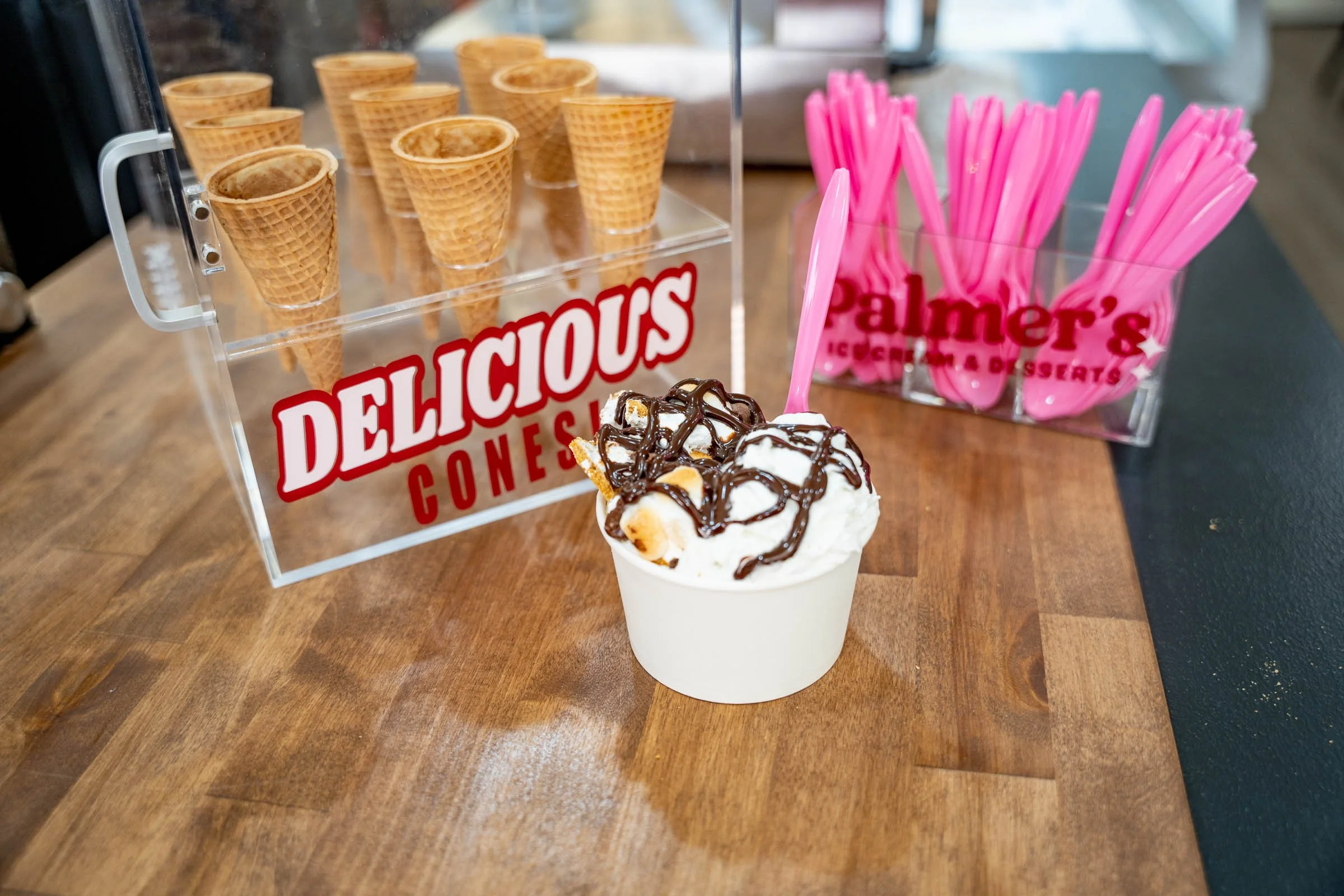 A cup of vanilla ice cream topped with chocolate syrup and marshmallows in the foreground, with waffle cones and pink spoons in containers in the background.