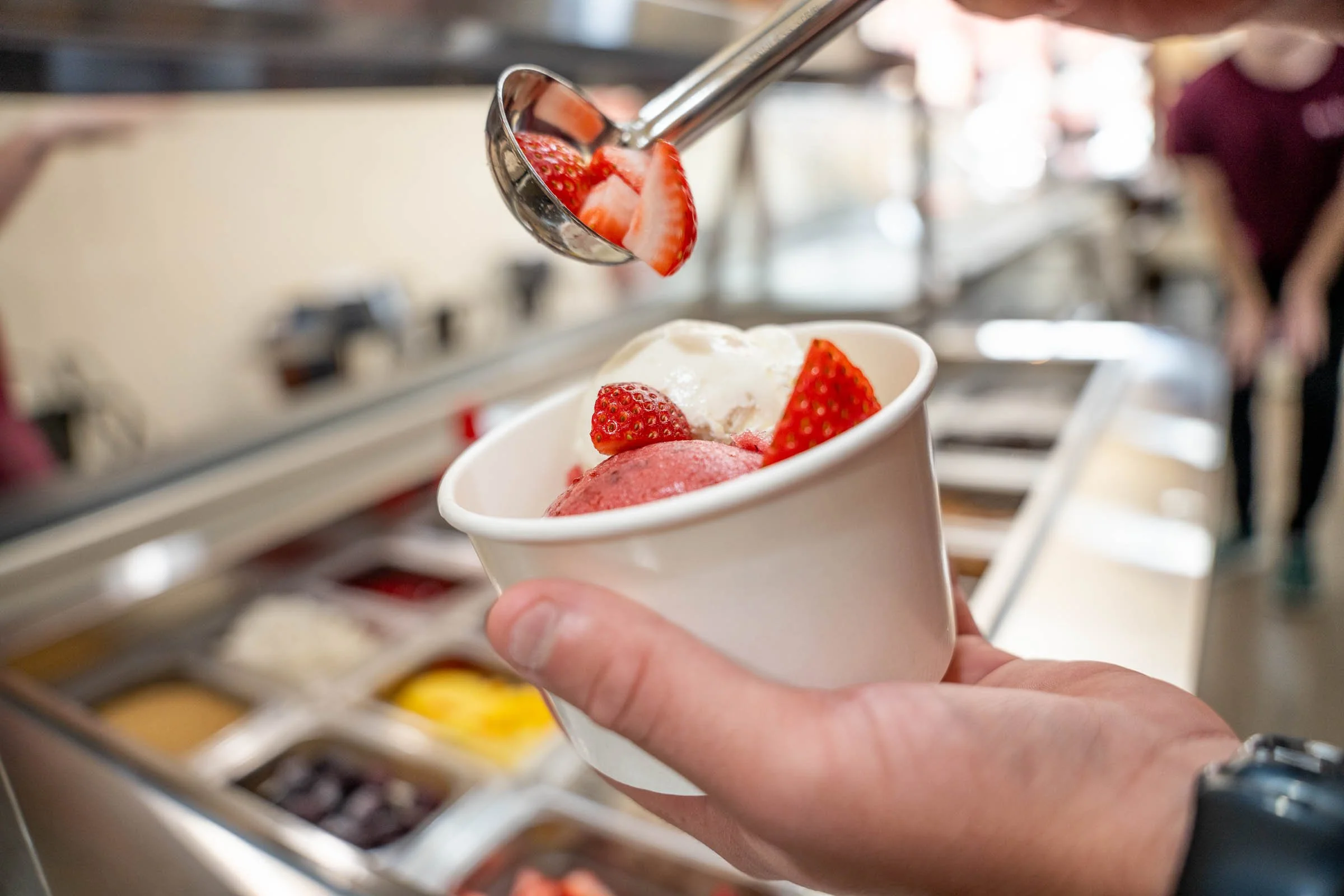 Close-up of a hand holding a small cup with strawberries and ice cream, with a scoop of strawberries being added using tongs at a dessert or ice cream shop.