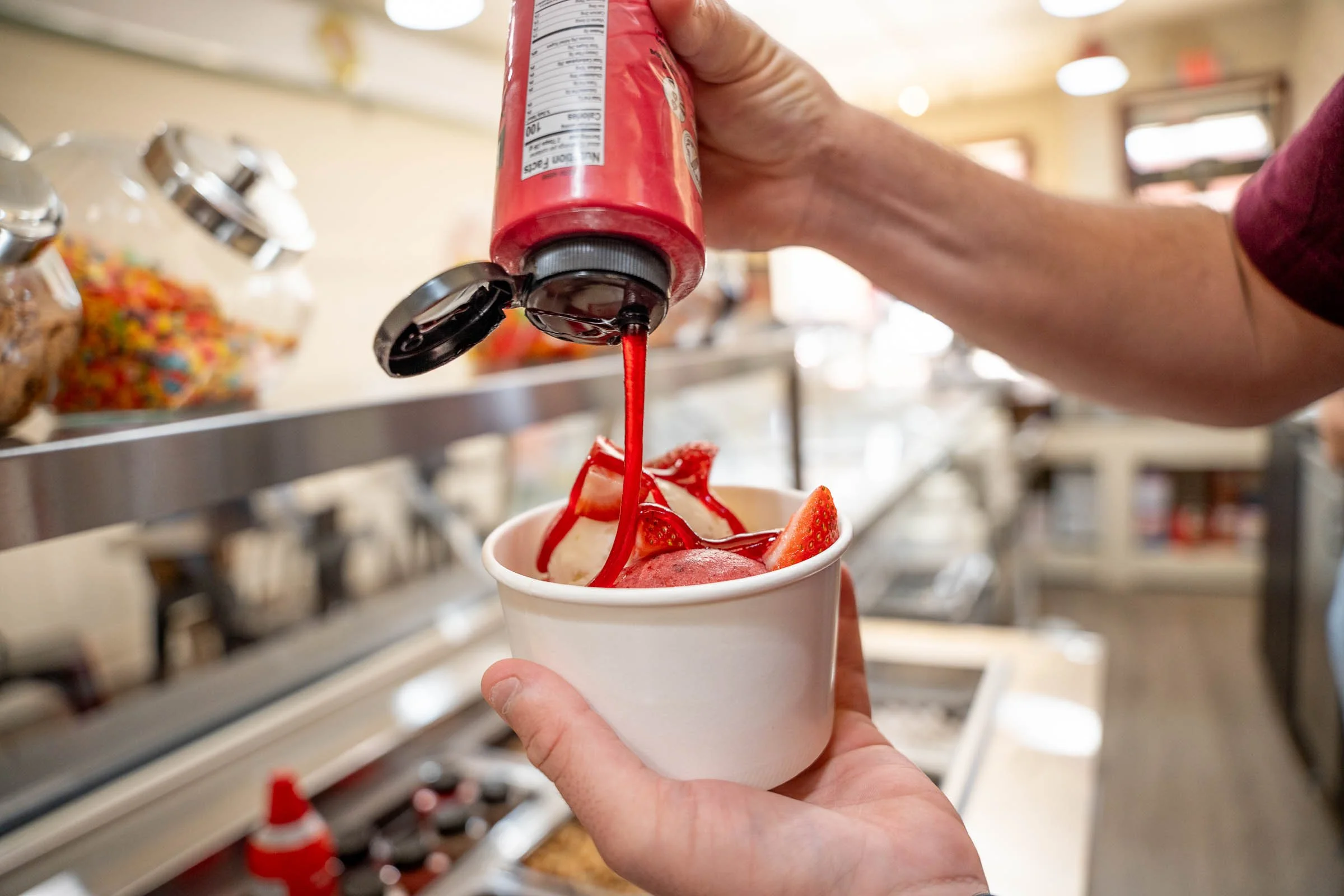 A person's hand holding a paper cup filled with vanilla ice cream topped with fresh strawberries and drizzled with strawberry syrup. A hand is pouring more strawberry syrup onto the ice cream from a red bottle.