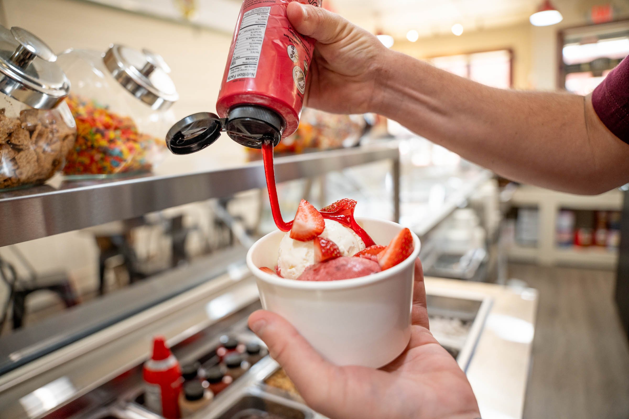 A person pouring red syrup onto a bowl of ice cream topped with strawberries.
