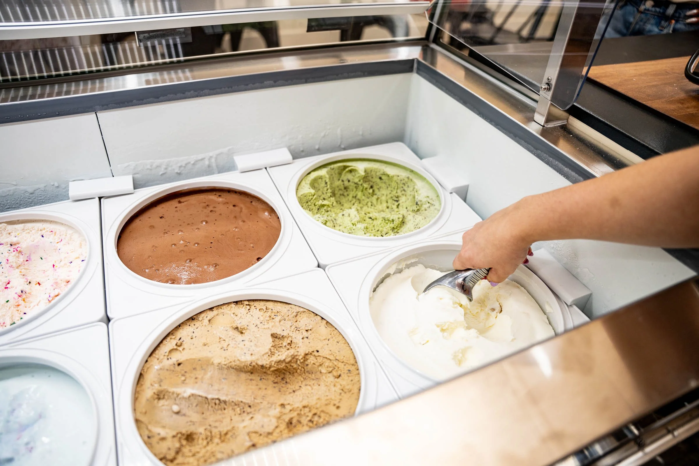 Assorted flavors of ice cream in a freezer, with a hand scooping vanilla ice cream.