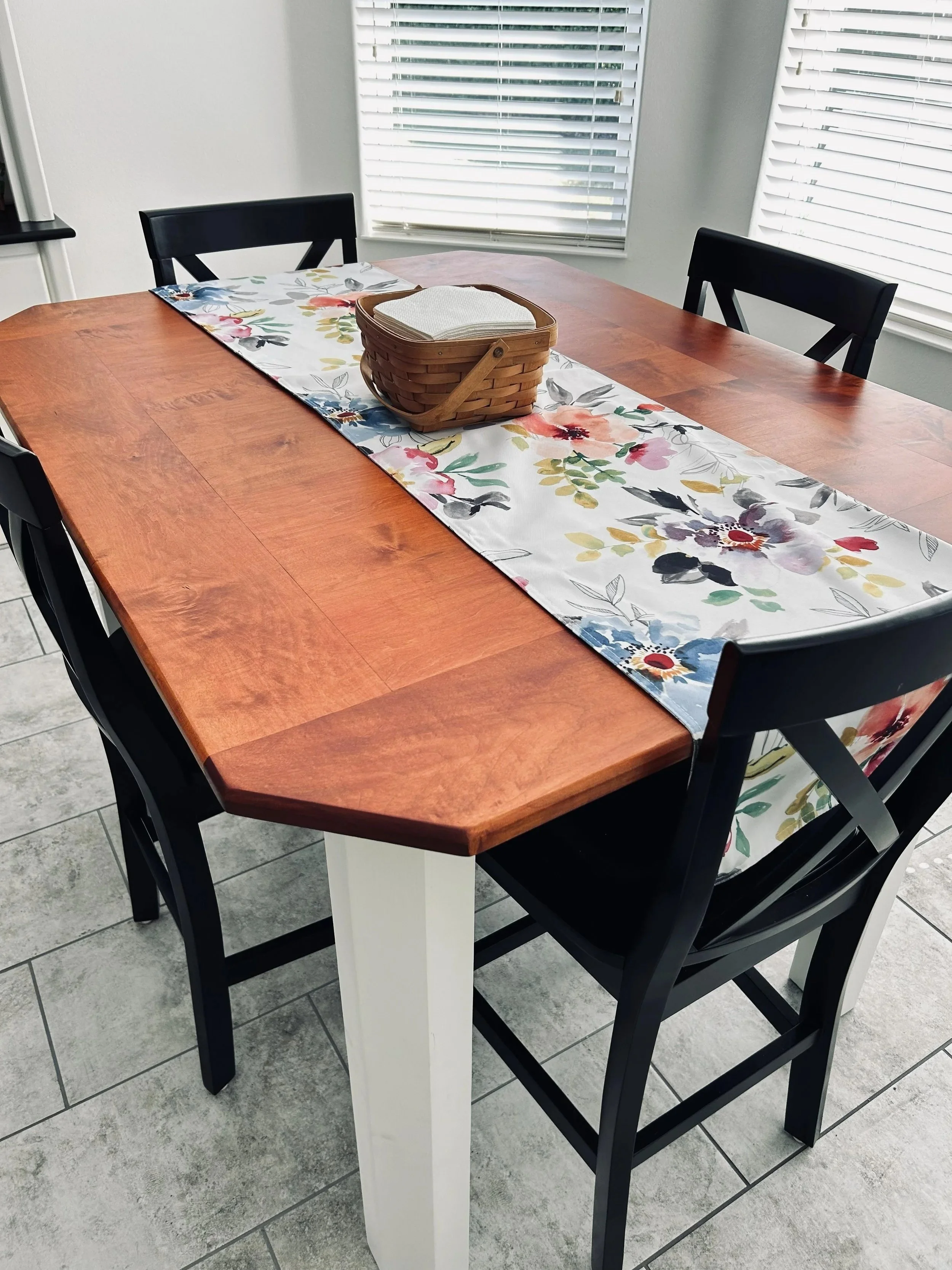 A wooden dining table with a floral table runner, black chairs, a basket with napkins in the center, and white walls with windows covered by blinds.