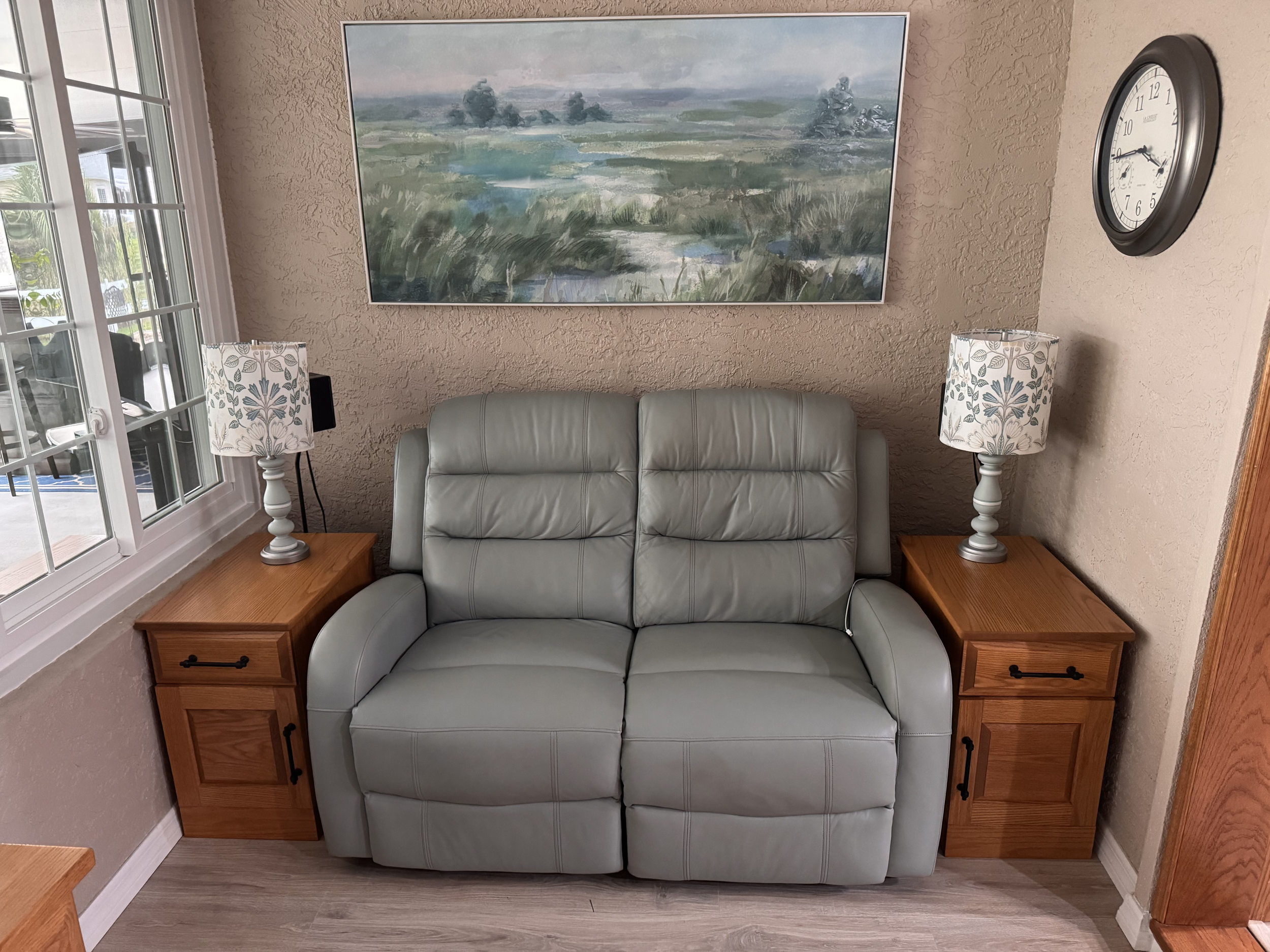 The image shows a living room corner with a light green leather sofa, two wooden side tables with matching table lamps featuring patterned shades, a large landscape painting hanging on a textured beige wall, a round wall clock, and a sliding glass door with a view of an outdoor patio.
