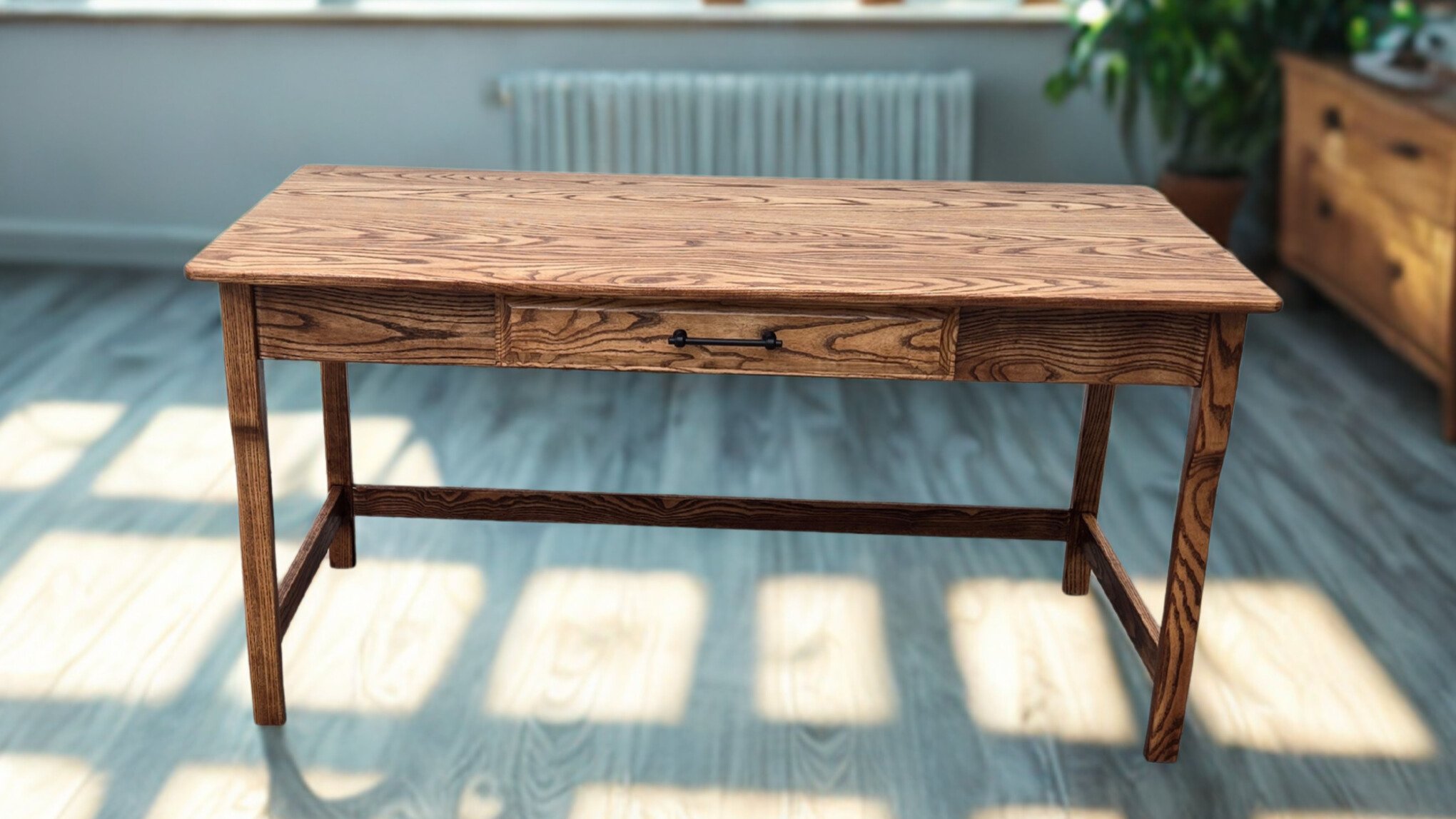A wooden table with a single drawer, situated in a room with sunlight casting shadows on the light-colored wooden floor. In the background, there is a gray wall, a radiator, and a green plant.