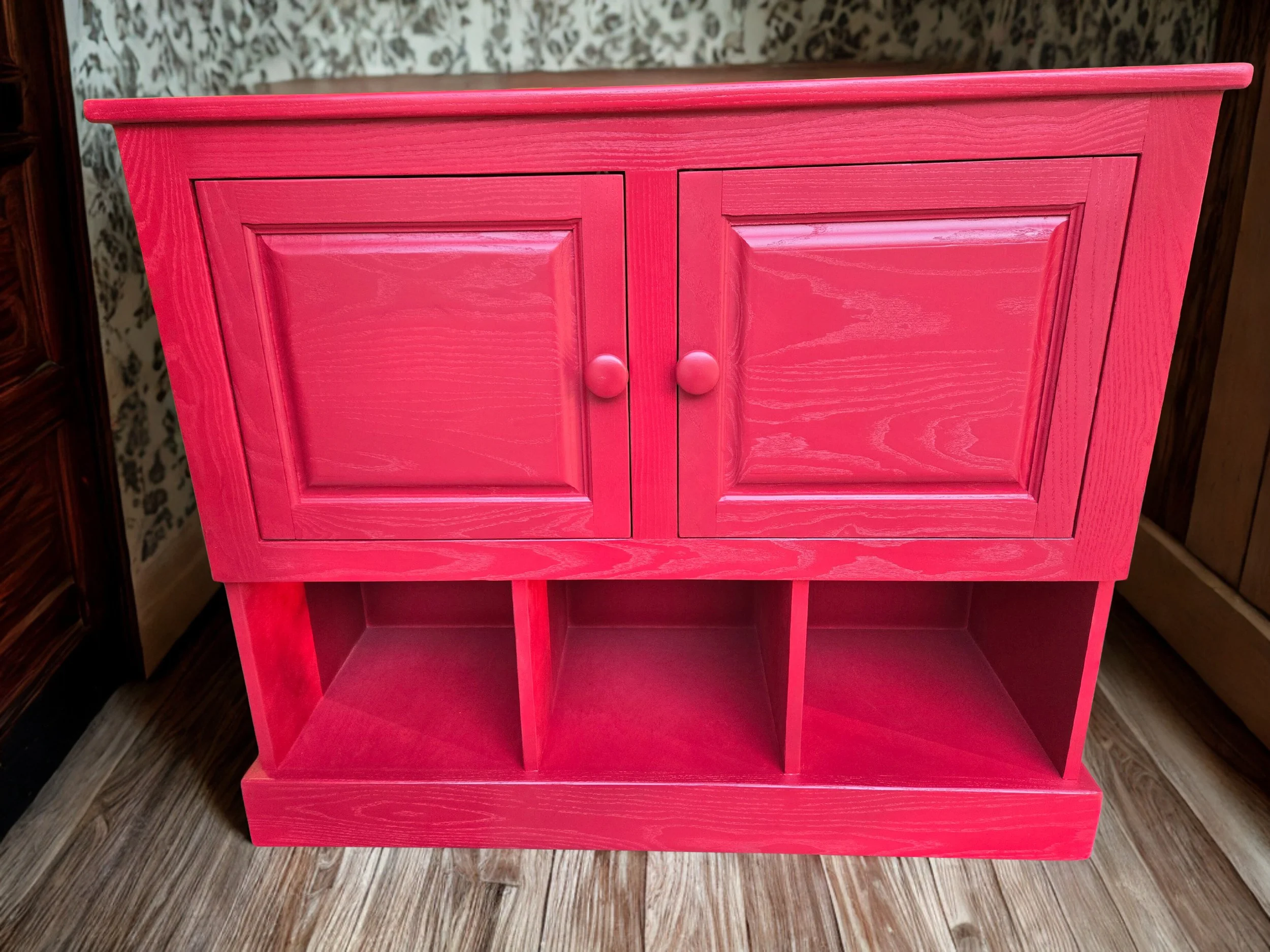 A bright pink wooden cabinet with two doors and round knobs, standing on a hardwood floor with a patterned wallpaper background.