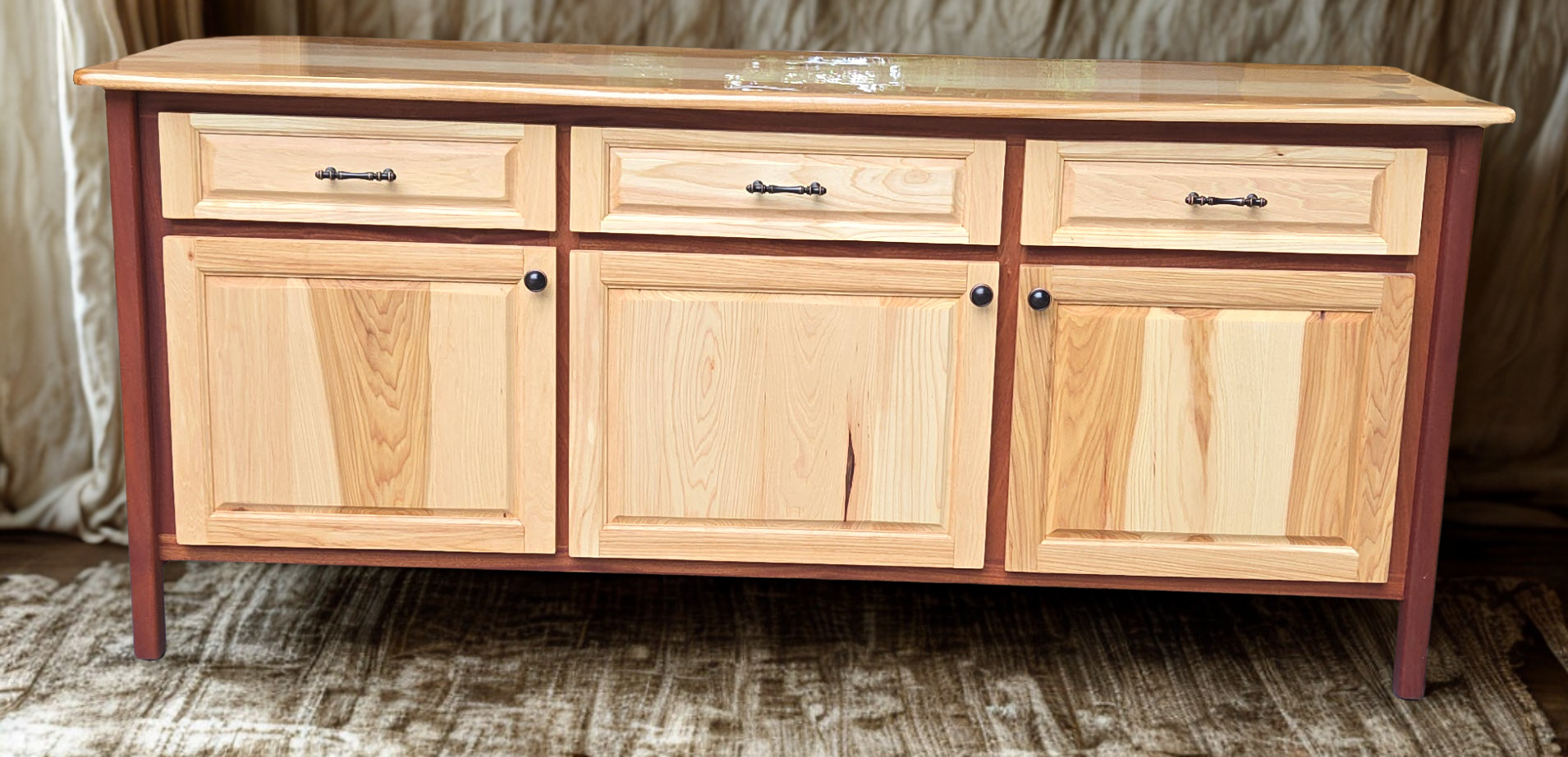 A wooden sideboard with a polished top, three small drawers at the top, and three large cabinets below, set on a patterned rug.