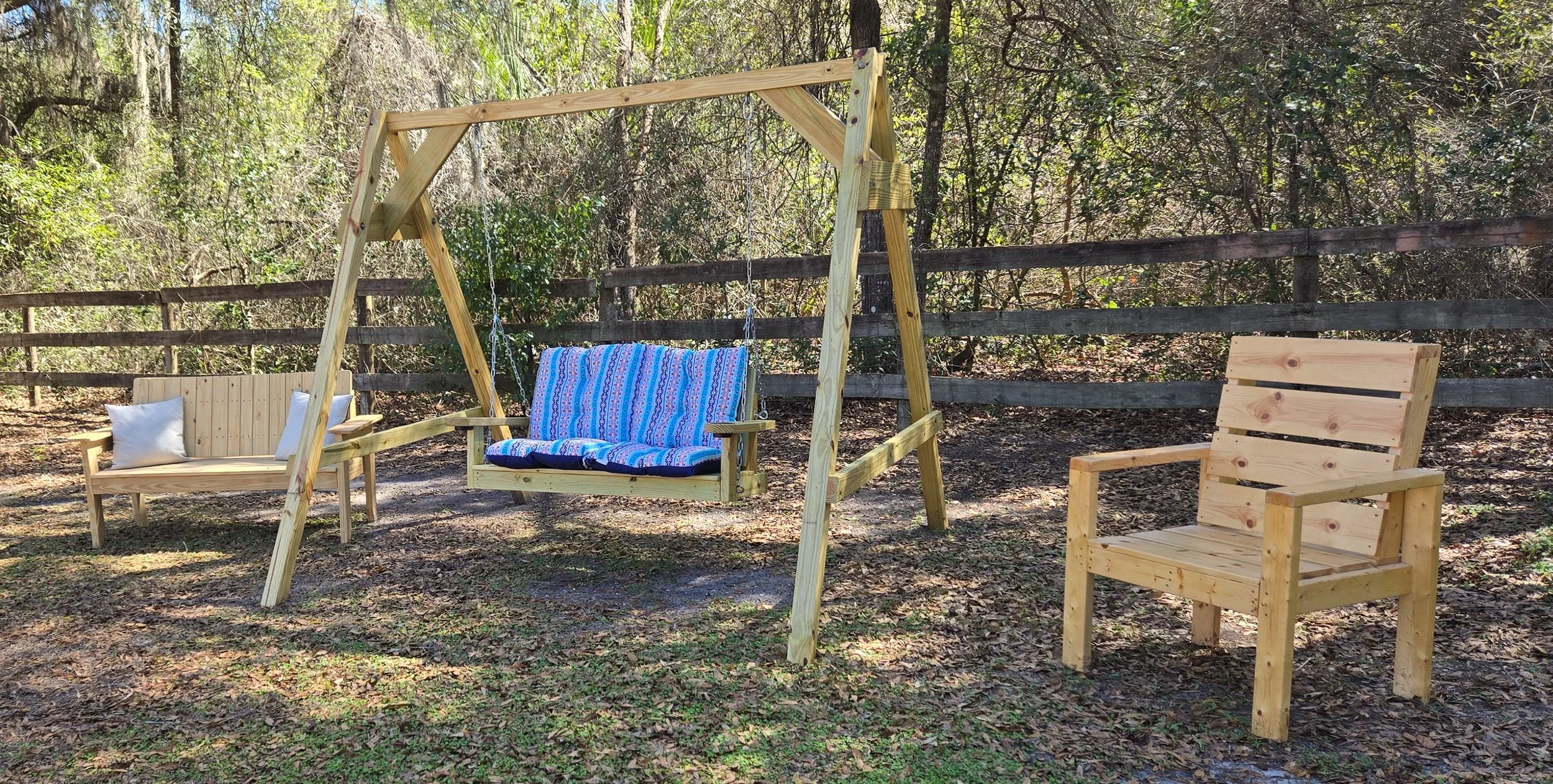 An outdoor scene with wooden furniture including a swing with a colorful blanket, a bench with white pillows, and an empty chair, set on dirt ground in a wooded area with trees and a fence in the background.