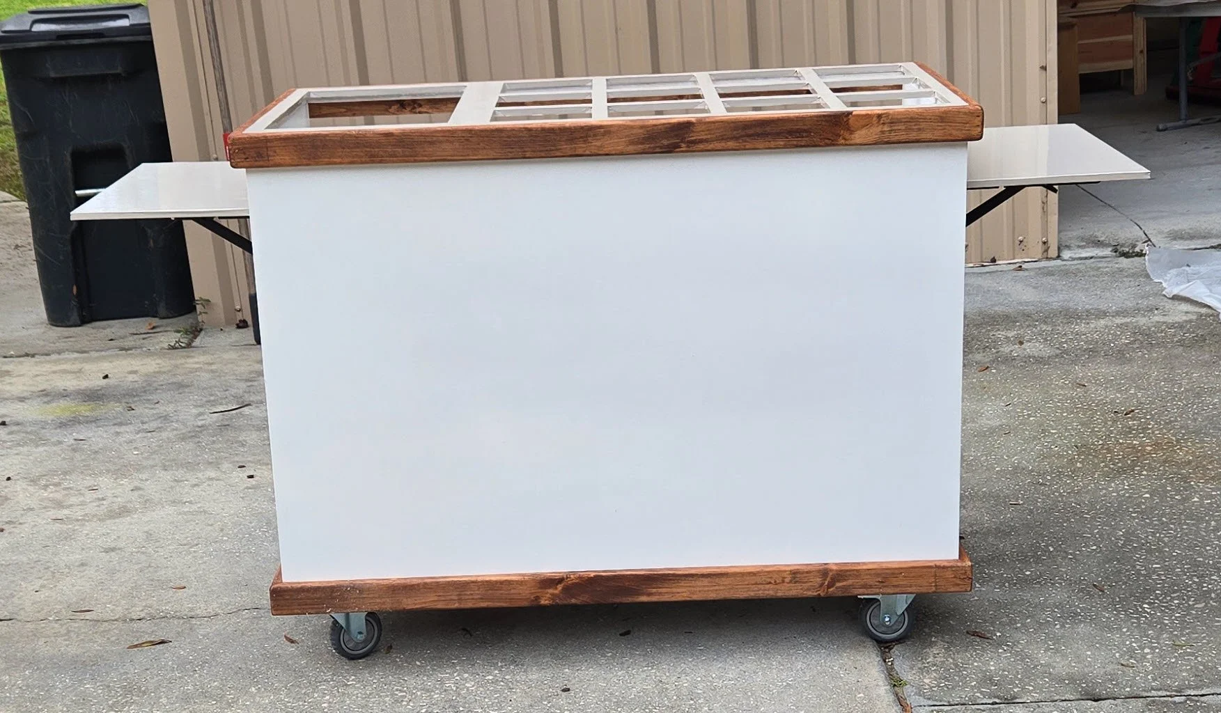 A mobile cart with a white body and wooden trim on top and bottom, on casters, positioned outdoors on concrete ground in front of a beige building.