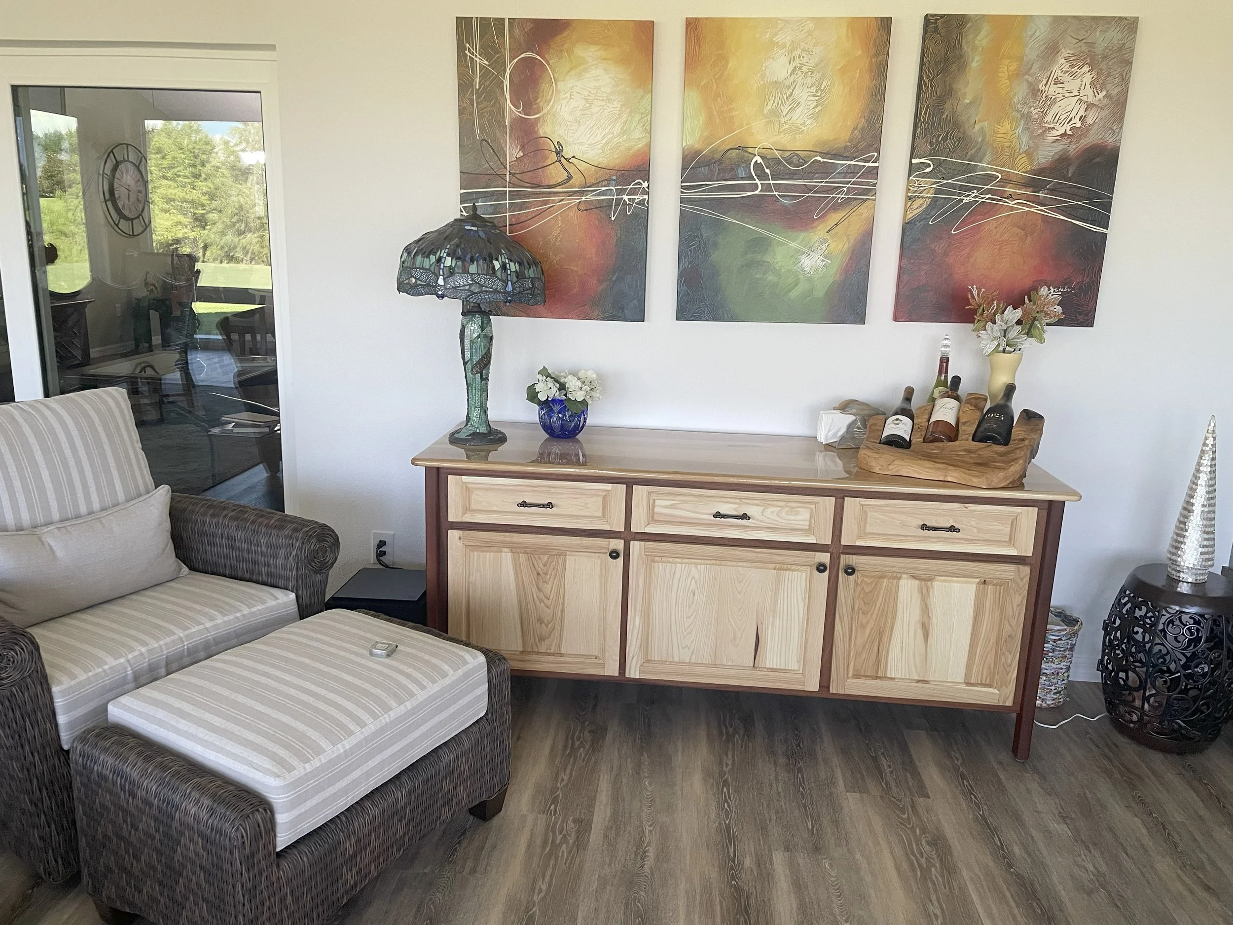 Living room with a wicker sofa with striped cushions, a wooden sideboard with artwork and decorative items, a glass lamp, and a sliding glass door revealing a green outdoor area.