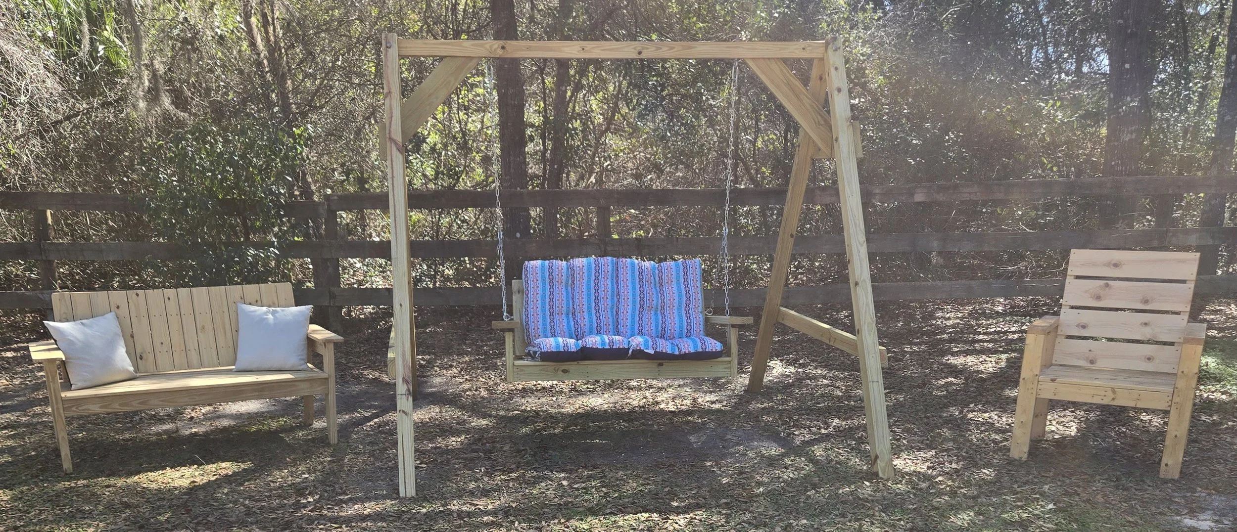 A wooden outdoor swing, bench, and chair arranged in a backyard garden with a wooden fence and trees in the background.