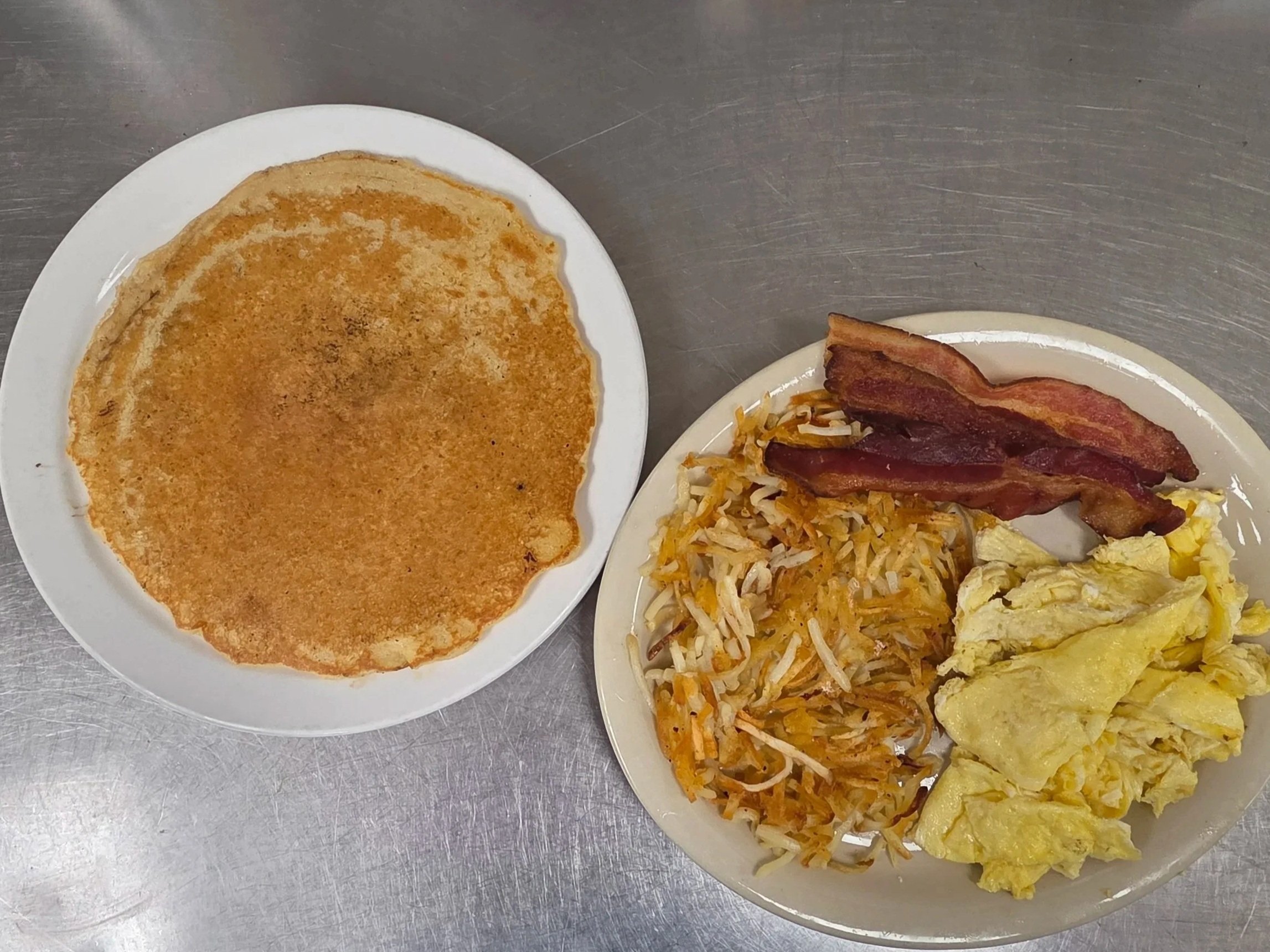 Two plates of breakfast food on a stainless steel table. The first plate has a large, round, golden-brown pancake. The second plate contains bacon, scrambled eggs, hash browns, and fried potatoes.
