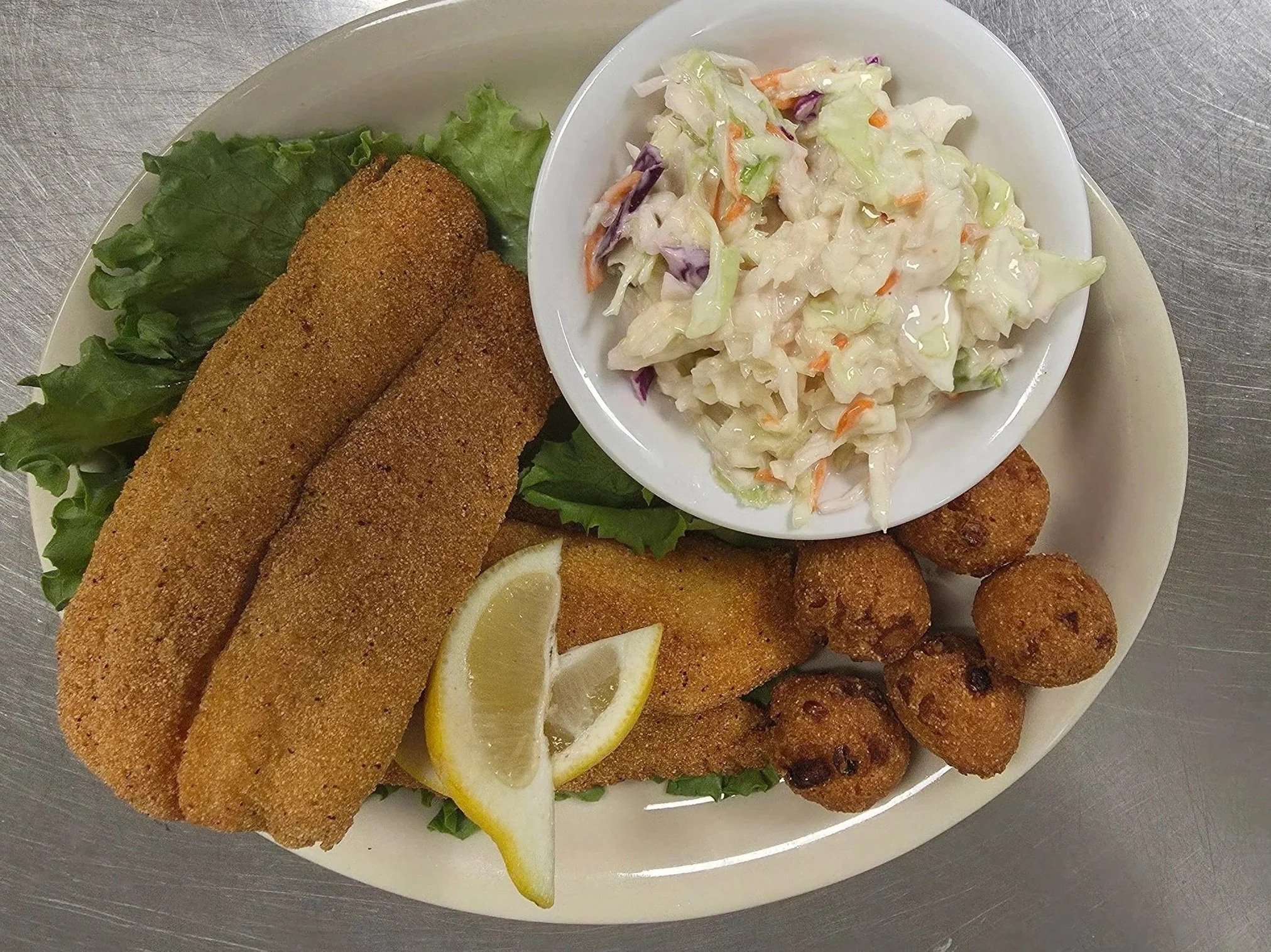 A white plate with fried fish fillets, lemon slices, and lettuce. The plate also has a bowl of coleslaw and some fried items along the edge.