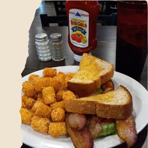 A plate with Buffalo chicken nuggets, two slices of toast, and bacon-wrapped green beans. In the background, there are salt and pepper shakers, a bottle of ketchup, and a red drink in a glass.