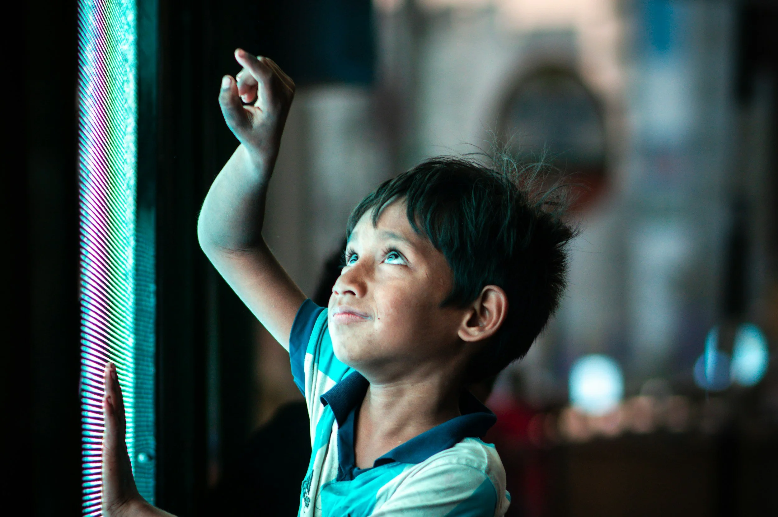 A young boy with dark hair and a striped shirt, looking up and reaching out to touch or interact with a colorful digital screen, standing in a dimly lit environment.