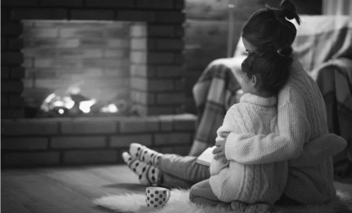 A woman and a girl sitting on the floor by a fireplace, hugging, with a mug in front of them.