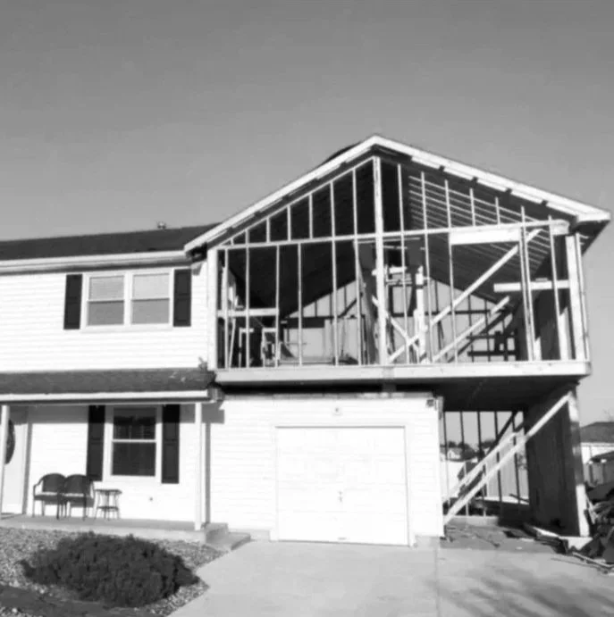 A two-story house with an unfinished glass-enclosed sunroom on the upper level, featuring visible wooden framing and construction materials.