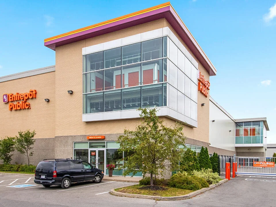 Exterior of an Entrepôt Public store, a large retail building with glass windows, beige walls, and orange signage. There are parked cars and a driveway leading to a gated entrance.