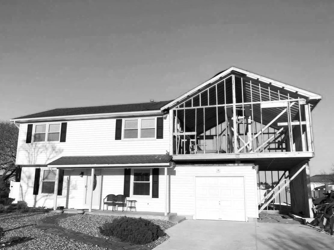 A two-story house with an unfinished second-story porch under construction, with visible wooden framing, and a garage on the first floor. The house has siding and black shutters, and the front yard has a porch with chairs and a small shrub.