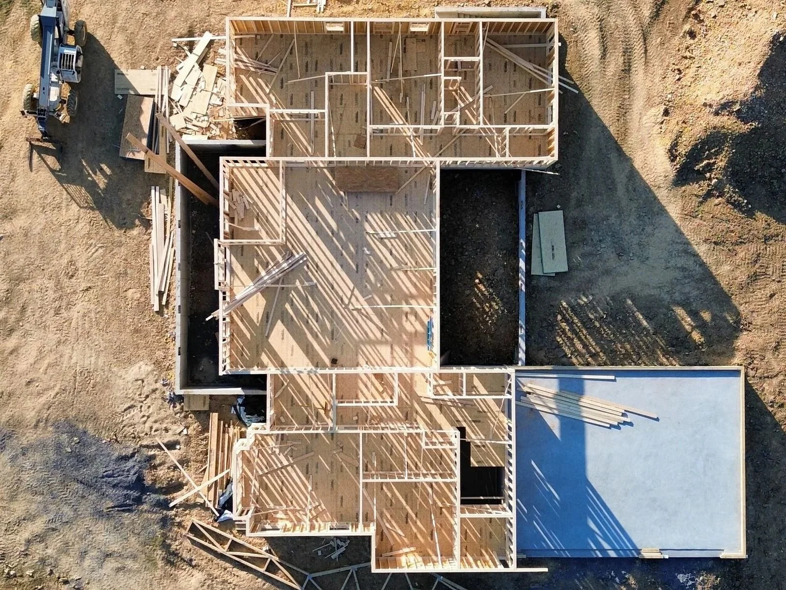 Aerial view of a house under construction showing its wooden framework, rooms, and shadows on the ground.