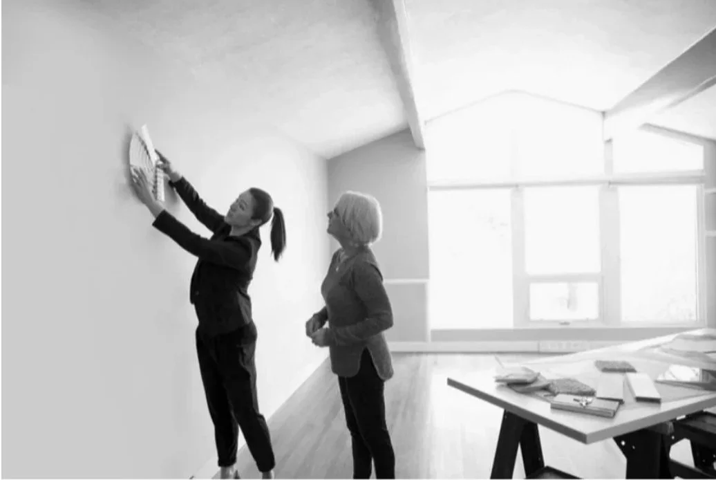 Two women cleaning a wall in a bright room with large windows, one is wiping the wall with a duster and the other is watching.