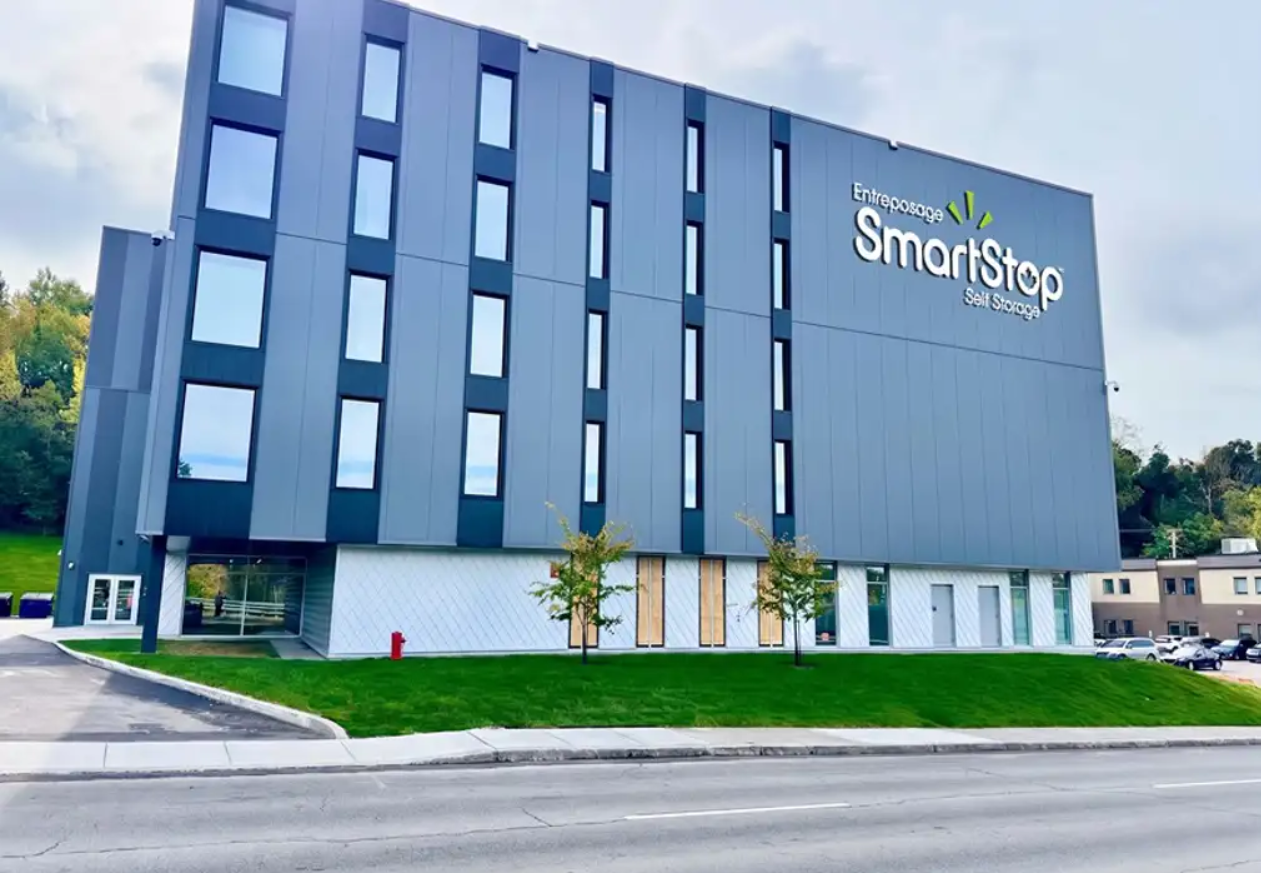 Exterior view of a multi-story self-storage building with a dark blue facade. The building features several narrow windows and a sign that reads "Entreposage SmartStop Self Storage." There are three small trees in front, a green lawn, and a parking area with several cars nearby.