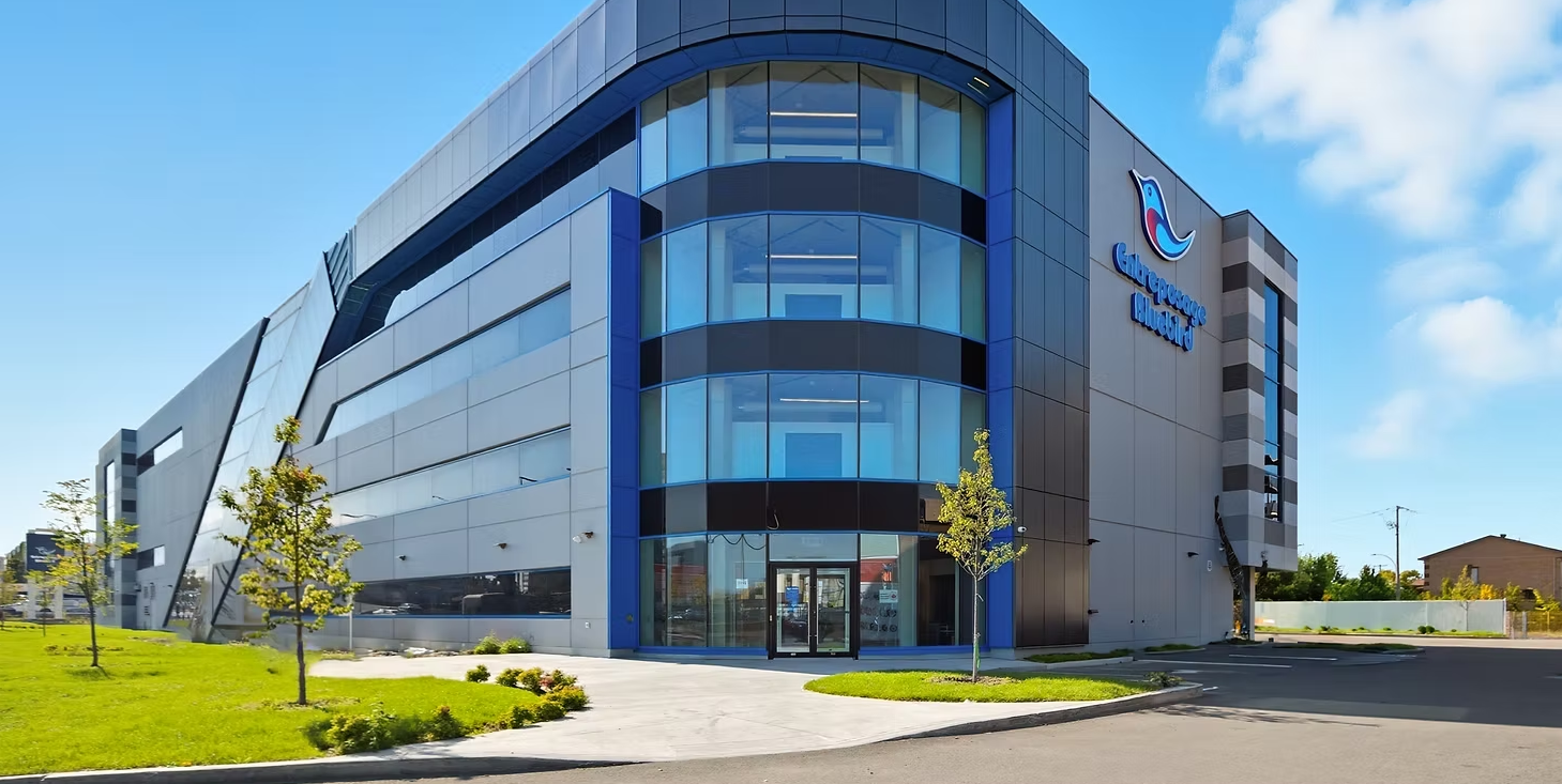 Modern multi-story office building with glass and metal exterior, blue accents, and the Canada Goose logo on the side.