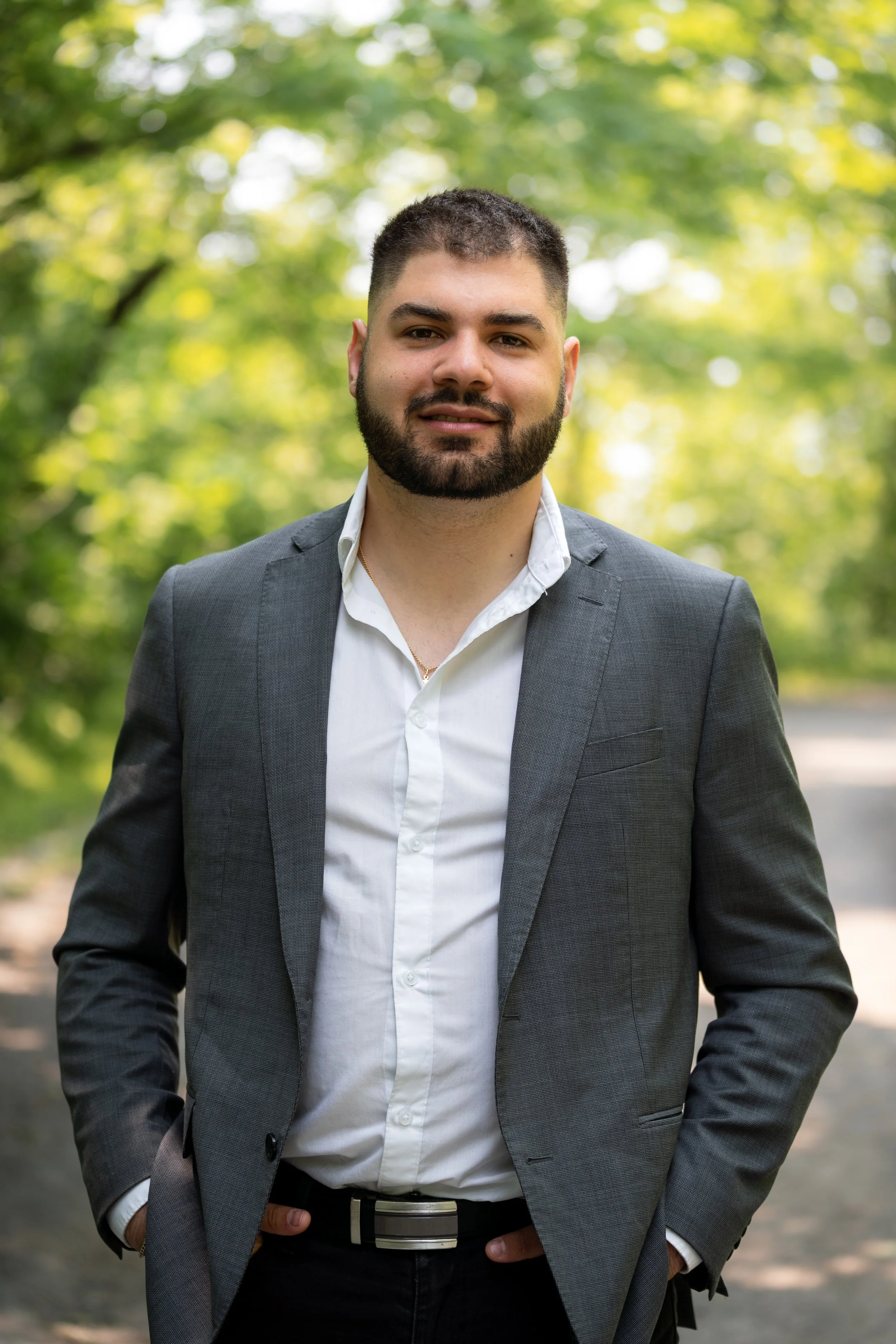 A man with a beard in a gray suit jacket and white shirt standing outdoors with trees and sunlight in the background.