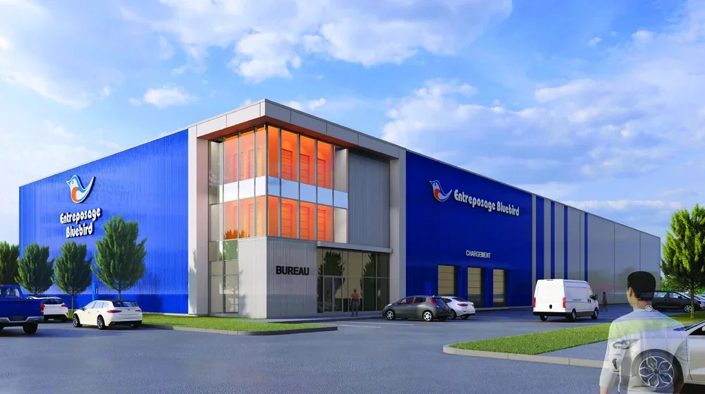 A modern industrial building with a blue exterior, large windows, and a sign that reads "Entrepots Bluebird." Several cars are parked outside, and a person is walking towards the entrance on a sunny day with some clouds in the sky.