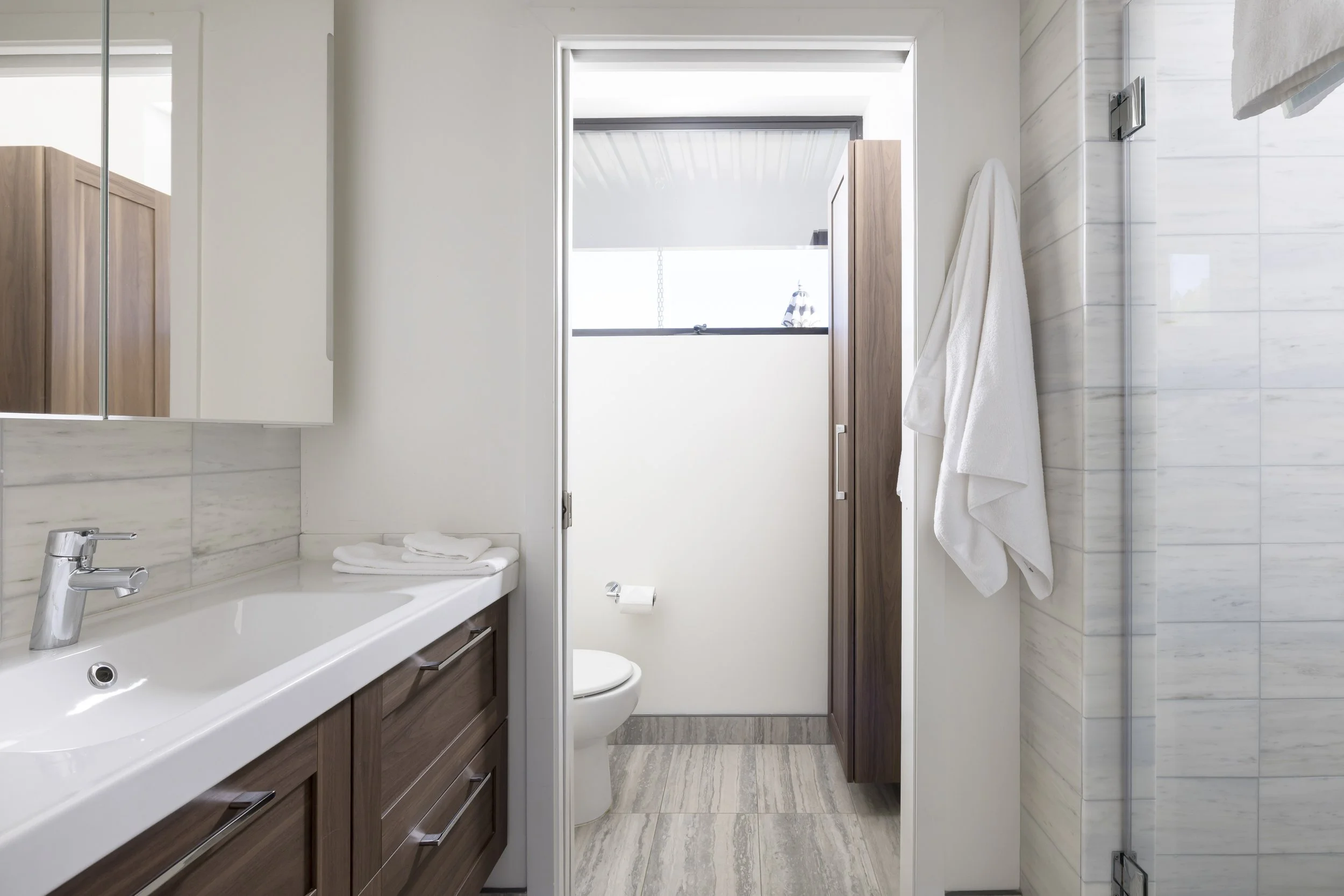 Modern bathroom with white sink, wooden cabinetry, toilet, and a window with a view of a life jacket hanging outside. Towel on hook and marble walls.