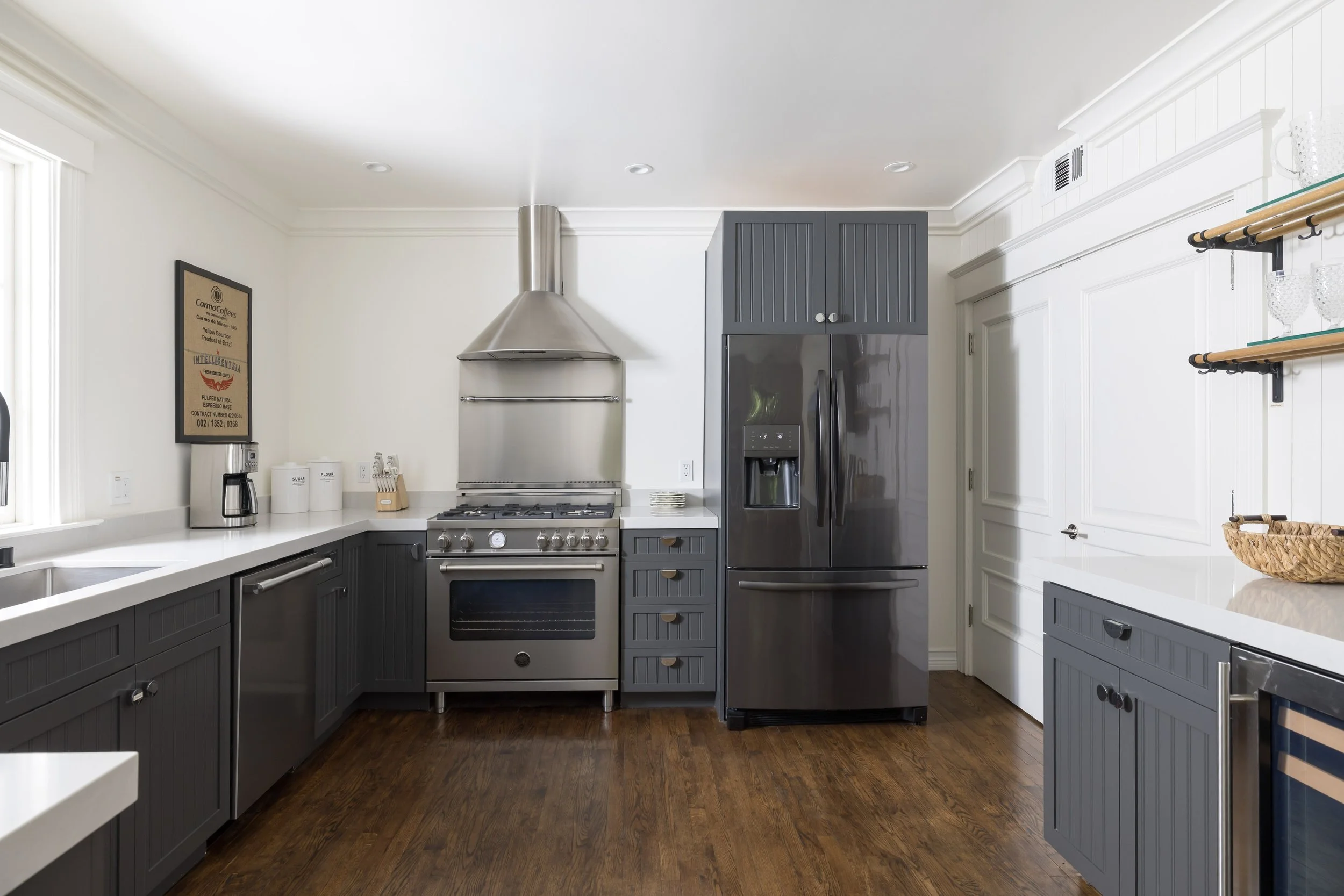 A modern kitchen with dark gray cabinets, stainless steel appliances, white countertops, and hardwood flooring. Visible items include a coffee maker, storage containers, and a refrigerator with water and ice dispenser.