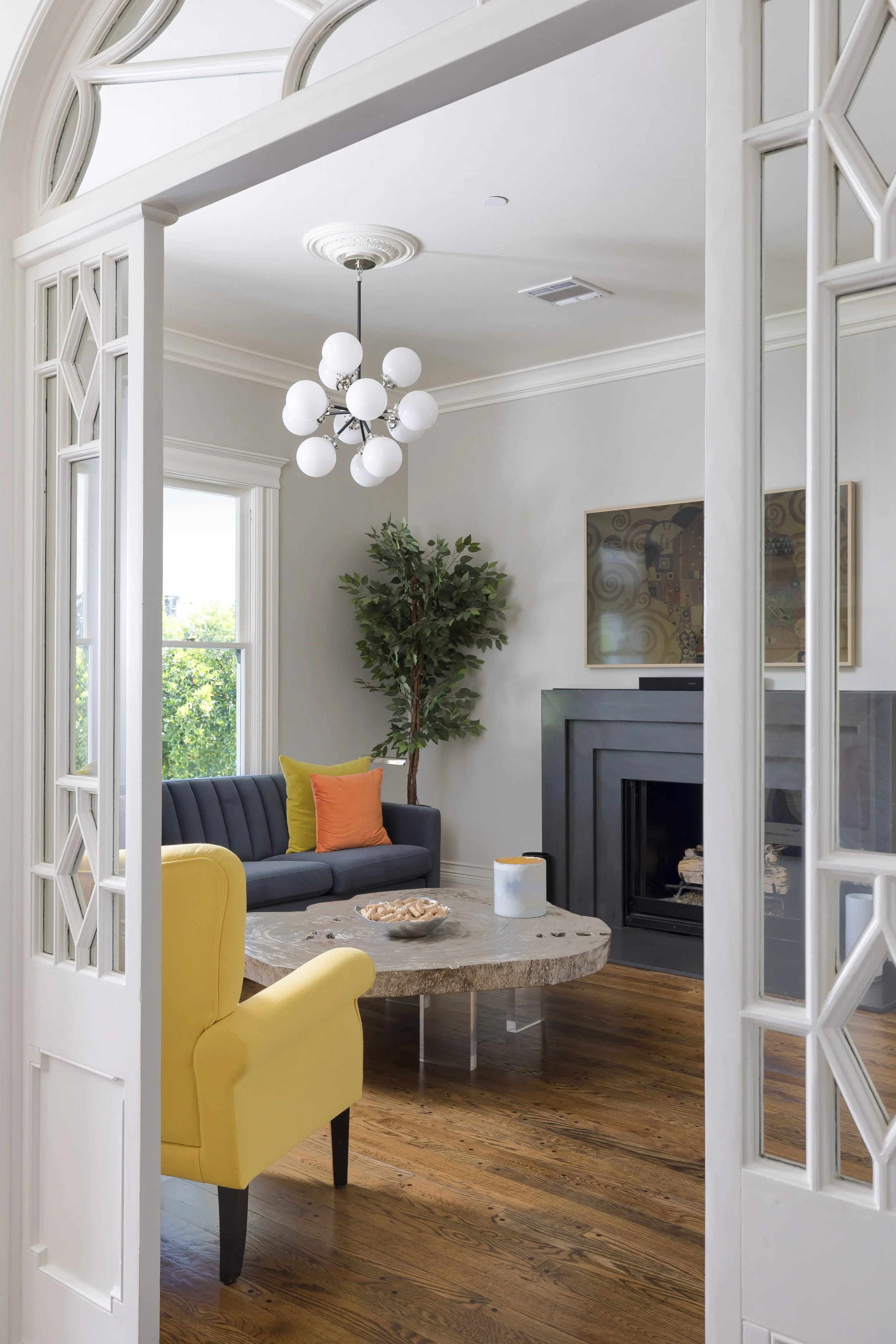 Living room with large windows, a black fireplace, a gray sofa with colorful pillows, a yellow armchair, a round wooden coffee table with a bowl of snacks, a tall potted plant, and a modern chandelier hanging from the ceiling.