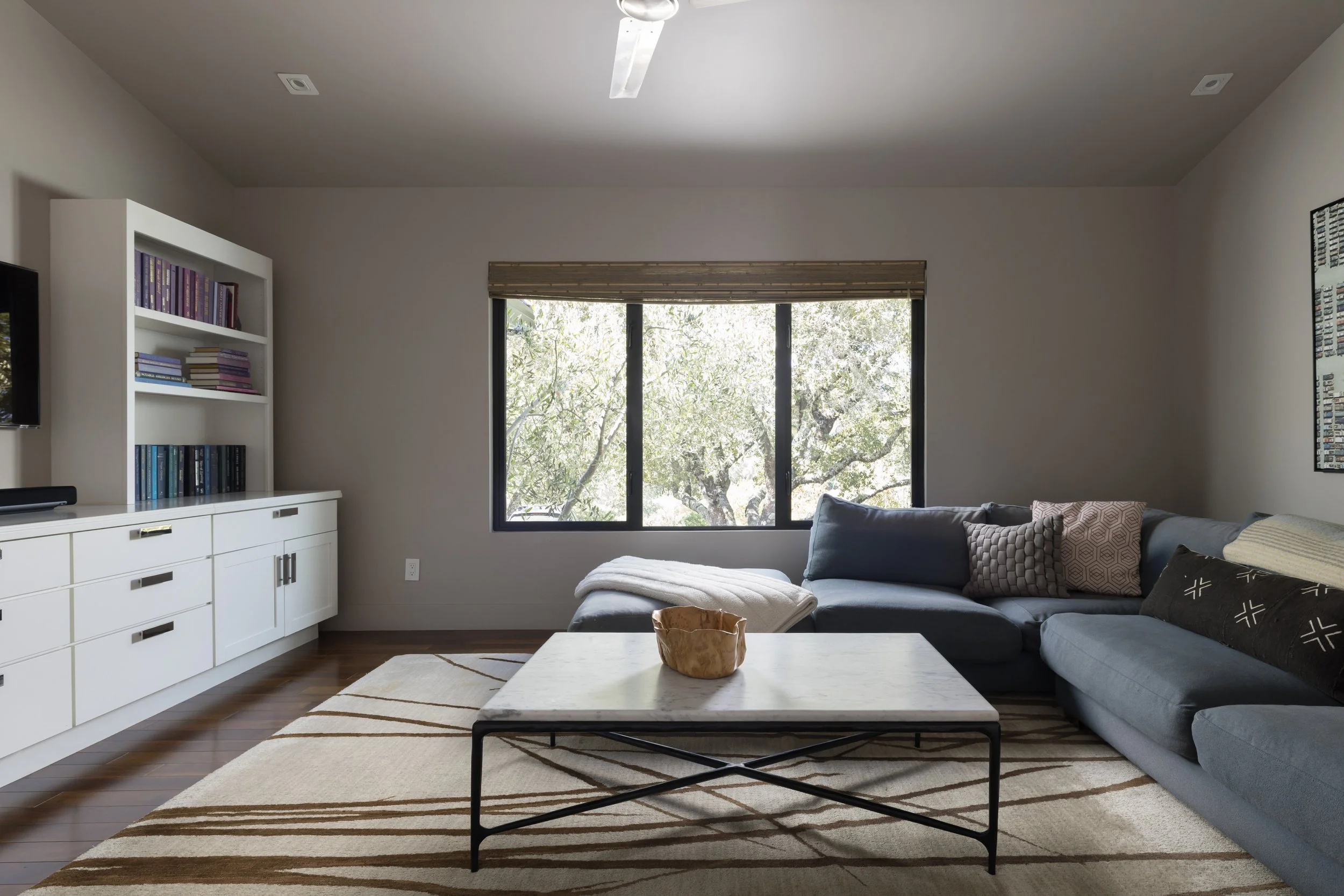 Living room with a sliding glass window, gray couch with patterned pillows, white bookshelf with books, wooden flooring, and a striped area rug.
