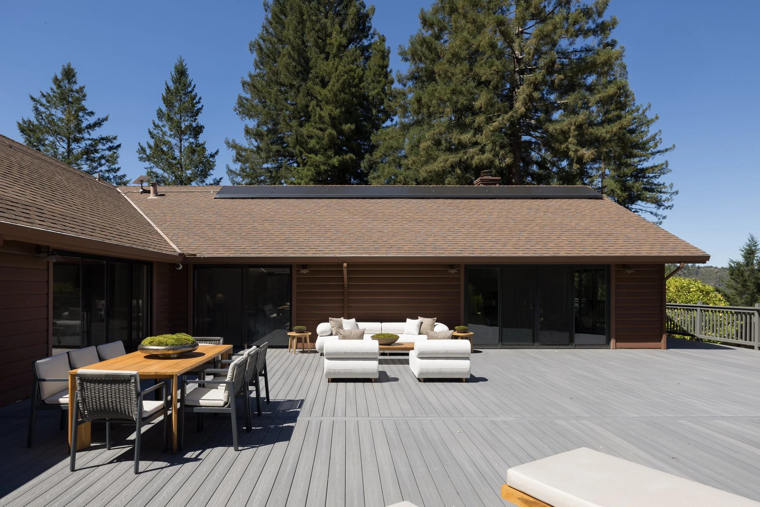 A spacious outdoor patio with grey wooden flooring, outdoor furniture including a white couch with multiple cushions, a wooden dining table with six chairs, and a small white bench, surrounded by a brown house with sliding glass doors, a deck railing