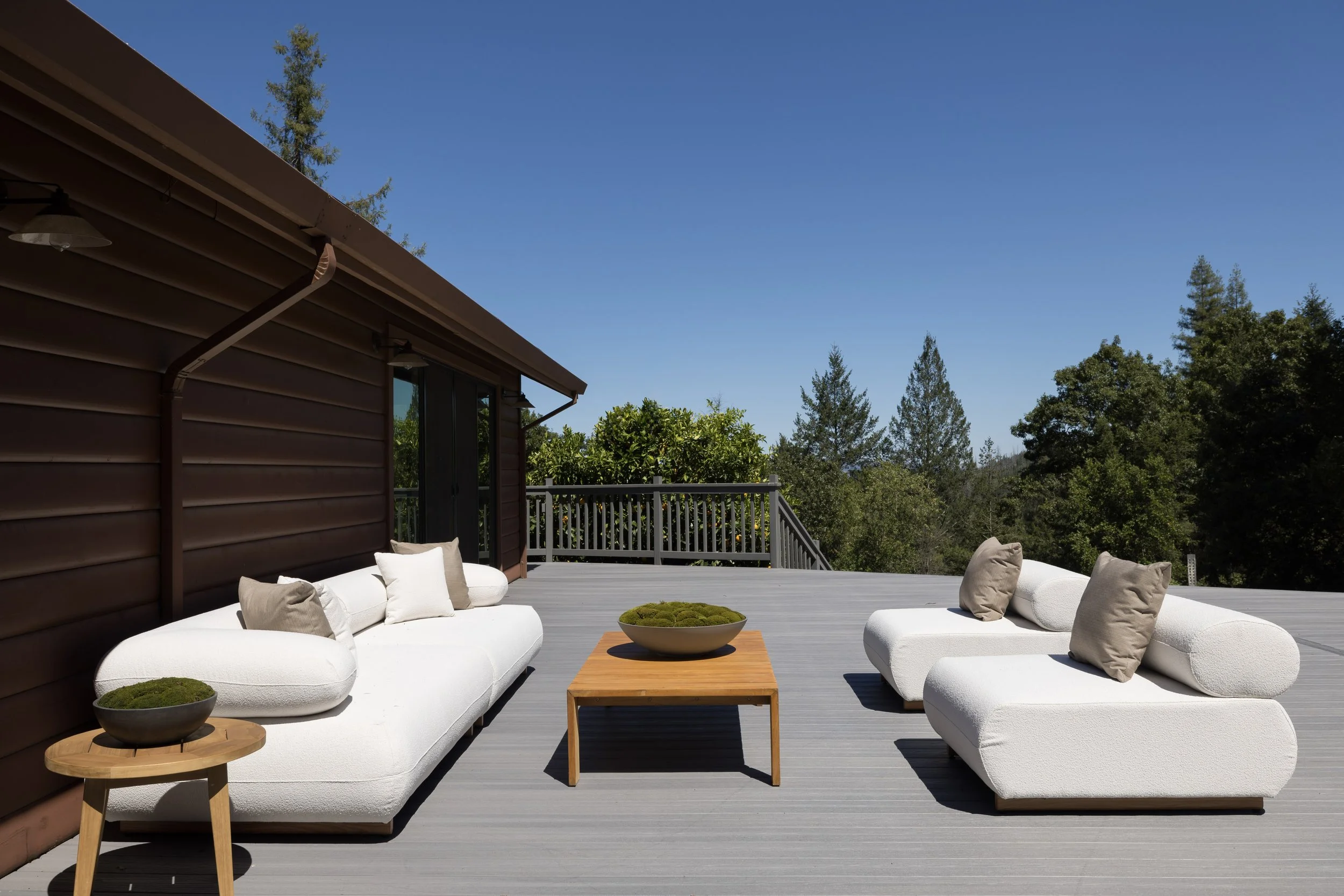An outdoor patio with modern white cushioned seating, a wooden coffee table, potted plants, and a railing, with trees and a clear blue sky in the background.