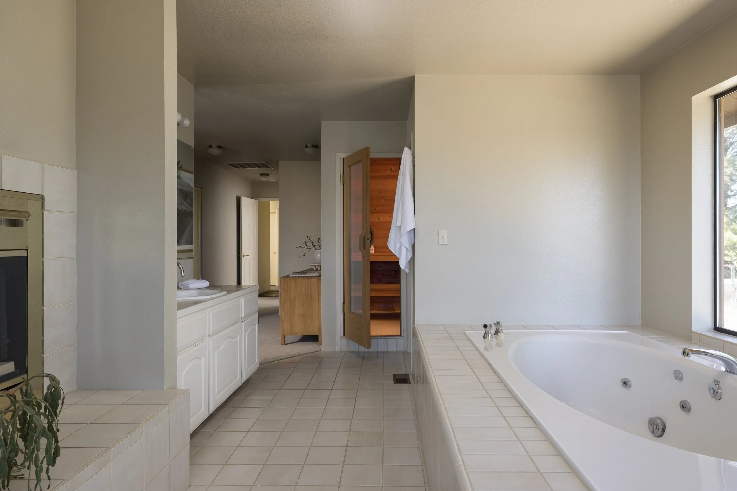 A spacious bathroom with a corner bathtub featuring multiple jets, beige tiled floor and ledge, large window on the right, with a white wall and a door leading to a sauna or heated space, and a sink area with towel and natural light.