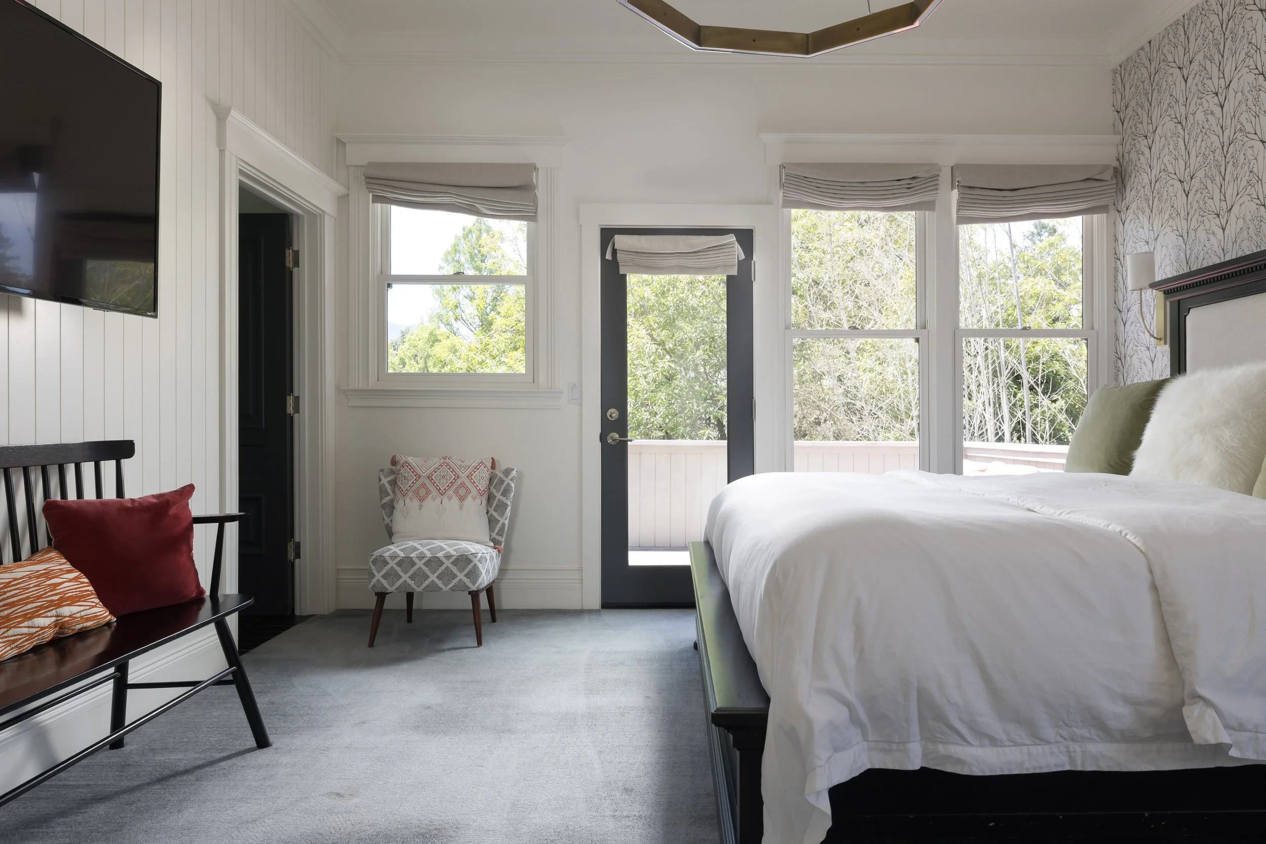 Bright bedroom with large windows showing green trees outside, a white bed with a dark frame, a patterned armchair, a black chair with red pillow, and a wall-mounted TV.