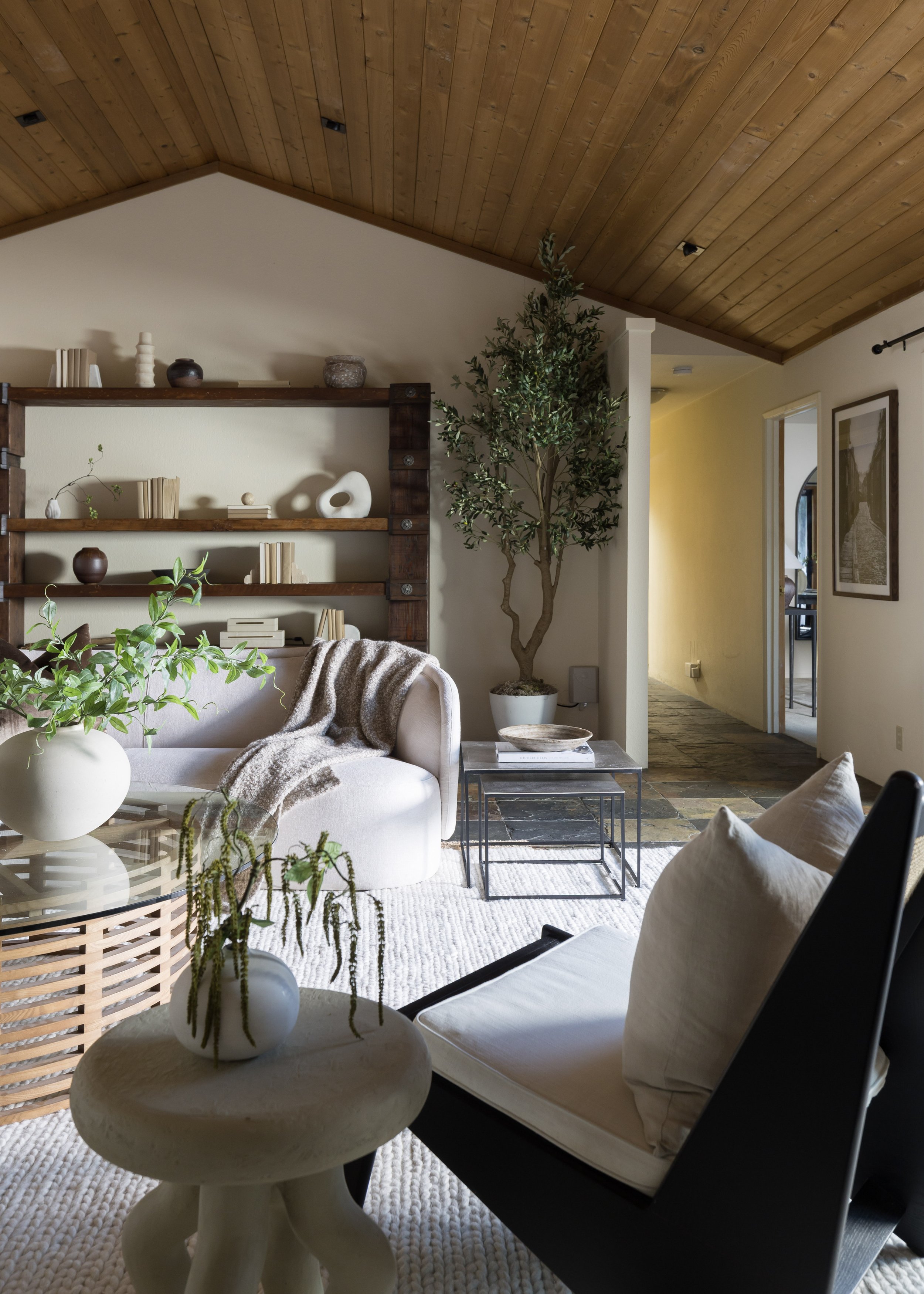 Living room with beige couch, black armchair, round side table with white vase and green plant, wooden ceiling, bookshelf with decorative items, large potted plant, and hallway with art on the wall.