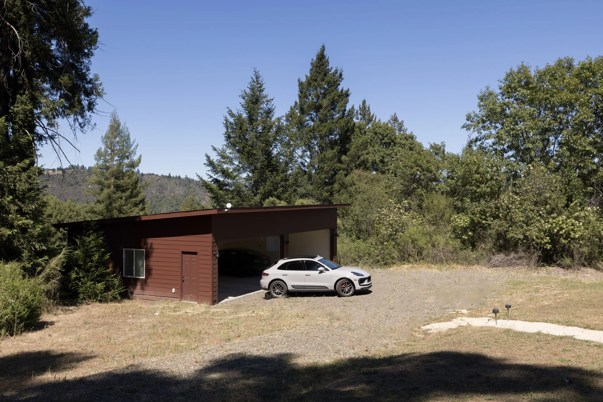 A white car parked in front of a red garage with trees and a mountain in the background under a clear blue sky.