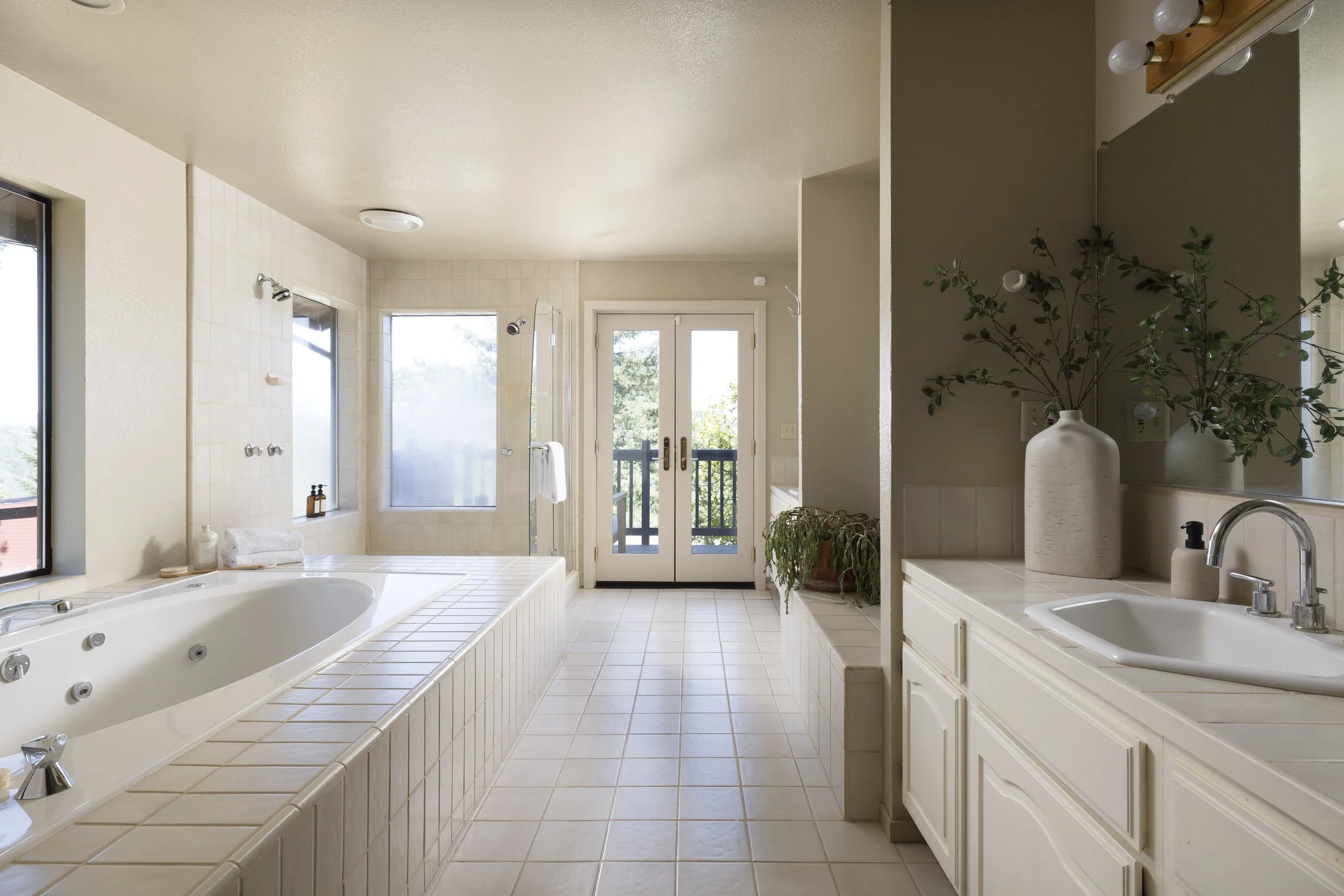 Bright bathroom with a jacuzzi tub, large windows, and a glass door leading to a balcony, decorated with plants and a mirror over the sink.
