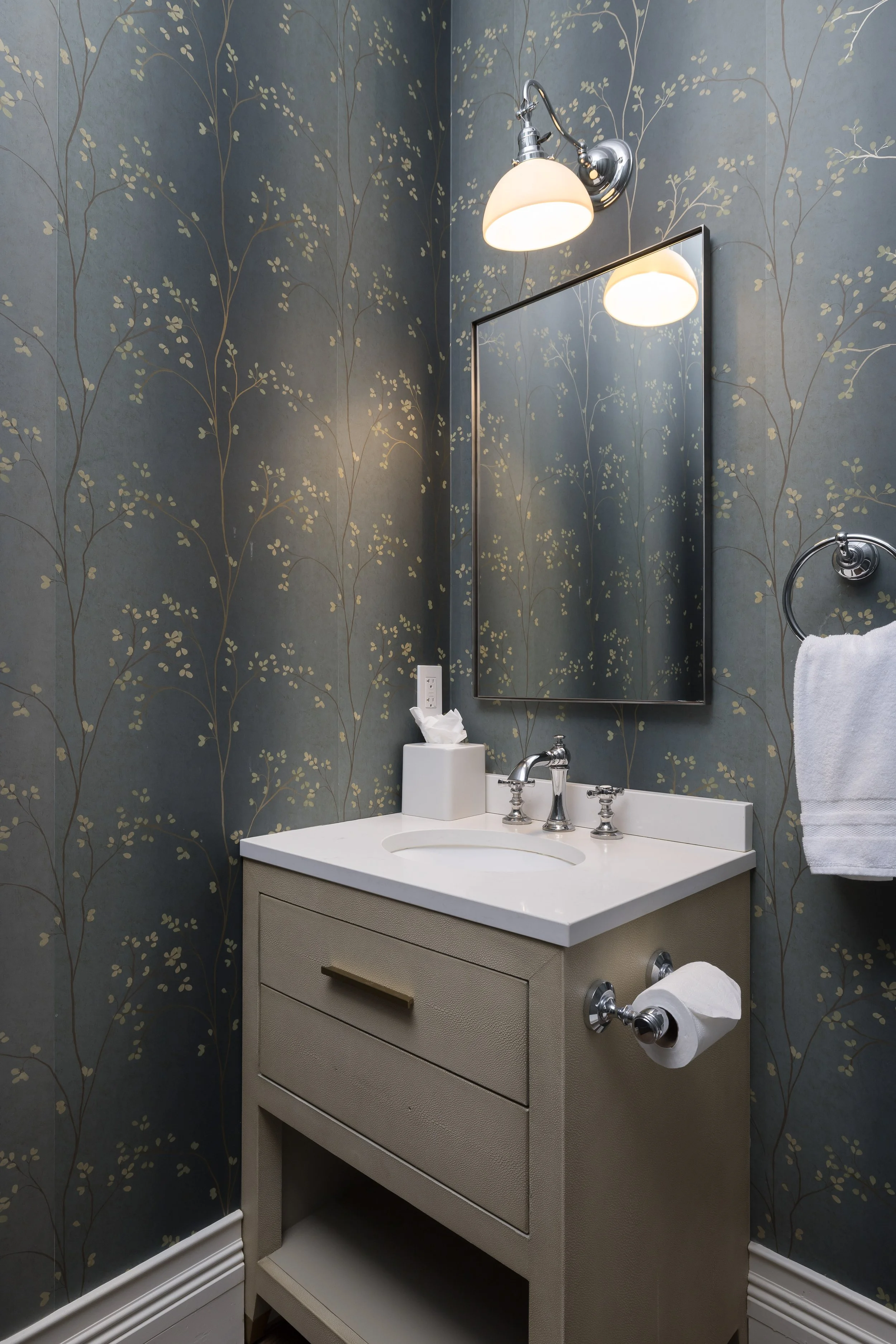 A bathroom sink vanity with a beige cabinet, a white countertop, a porcelain sink, and a silver faucet. There is a tissue box on the counter and a hand towel on a ring. The wall behind has blue wallpaper with a gold floral pattern. A rectangular mirr